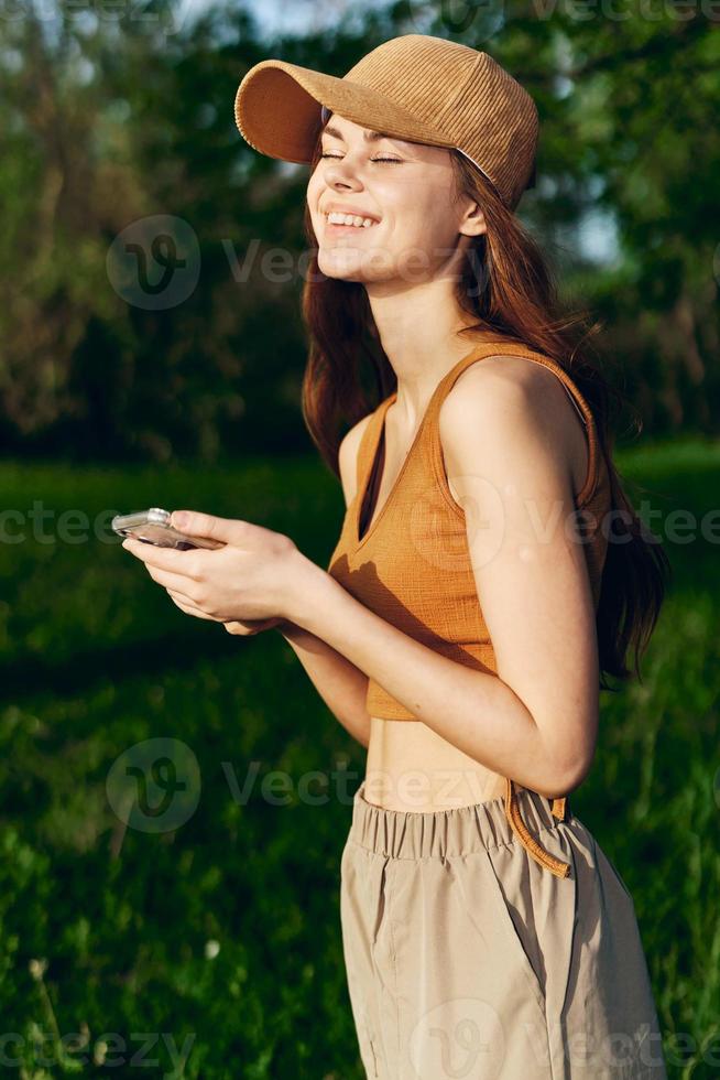 mujer blogger con teléfono en mano en naturaleza en contra un fondo de verdor sonriente en el Brillo Solar vistiendo un gorra después hacer ejercicio foto