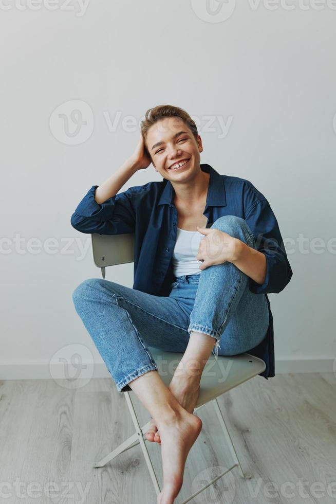 un joven mujer sentado en un silla a hogar sonriente con dientes con un corto Corte de pelo en pantalones y un mezclilla camisa en un blanco antecedentes. niña natural poses con No filtros foto