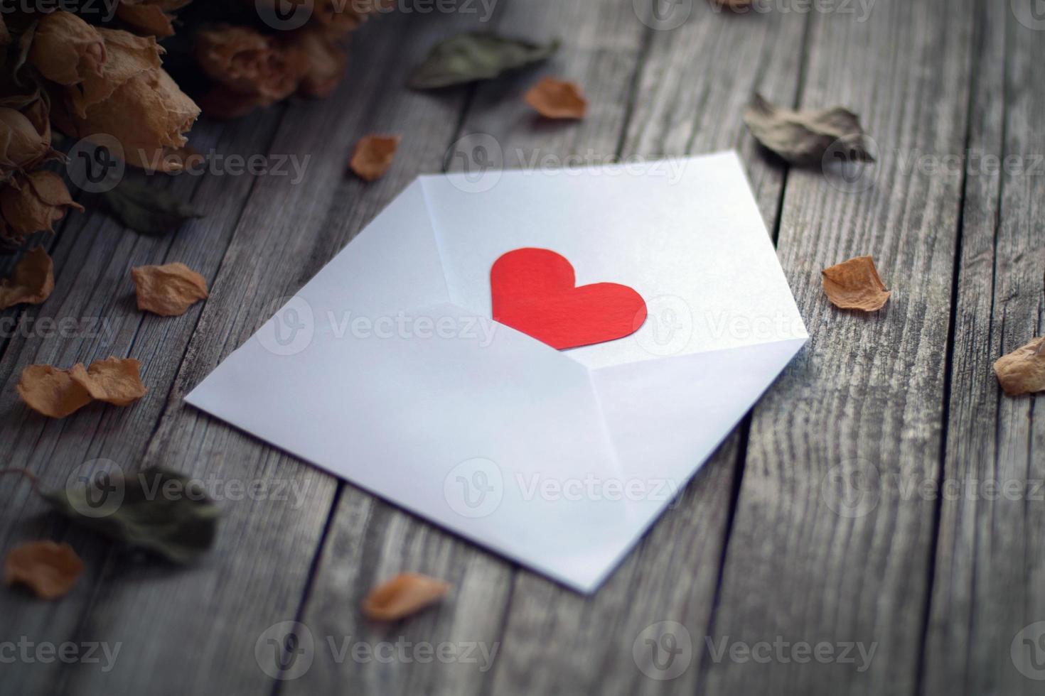 Open envelope with single red paper heart framed by dry roses on photo
