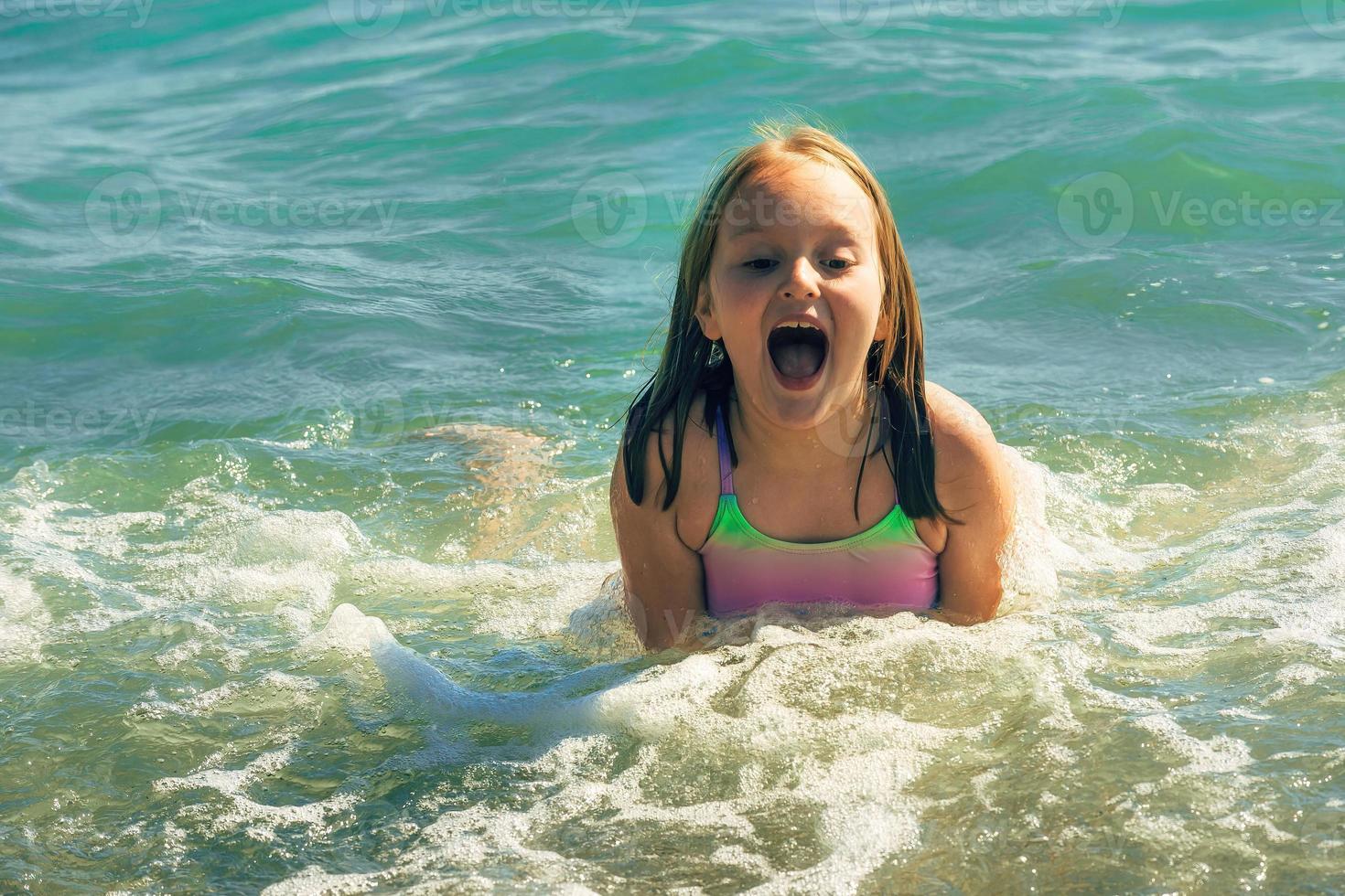 un pequeño niña en un traje de baño nada alegremente en el mar en un calentar verano soleado día foto