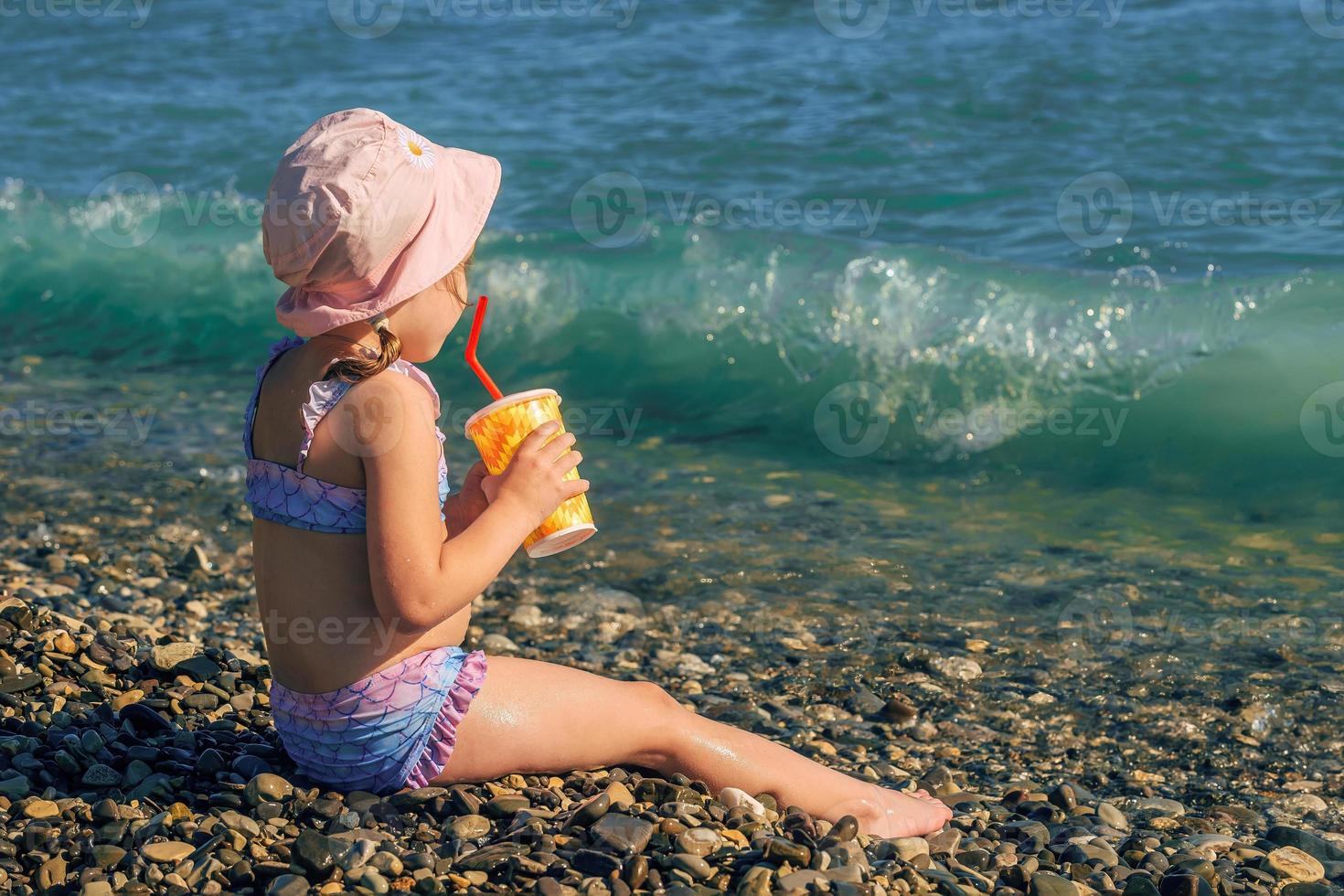 a little girl in a bathing suit and panama is sitting on the seashore, in her hands is a glass with a straw and a summer refreshing cocktail photo