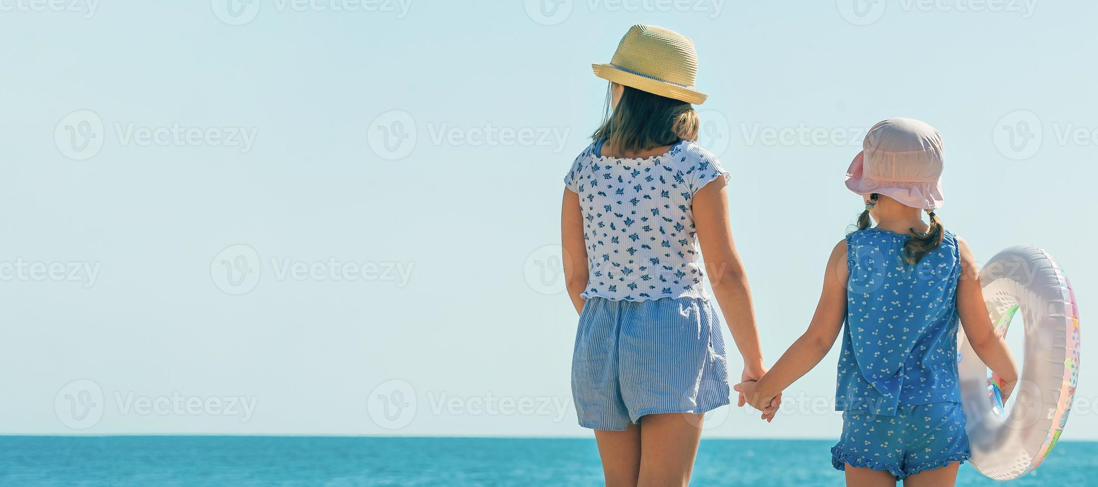 two little girls against the background of the blue sky and the ocean, they are dressed in summer beach clothes, in panama hats and with an inflatable swimming ring, banner, copy space photo