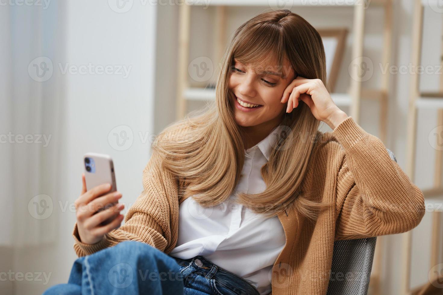 Cute young blonde lady in warm sweater doing video call with friends use phone sitting in armchair at modern home interior. Pause from work, take a break, social media in free time concept. Copy space photo