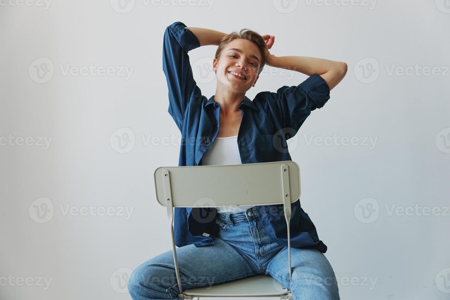 un joven mujer sentado en un silla a hogar sonriente con dientes con un corto Corte de pelo en pantalones y un mezclilla camisa en un blanco antecedentes. niña natural poses con No filtros foto