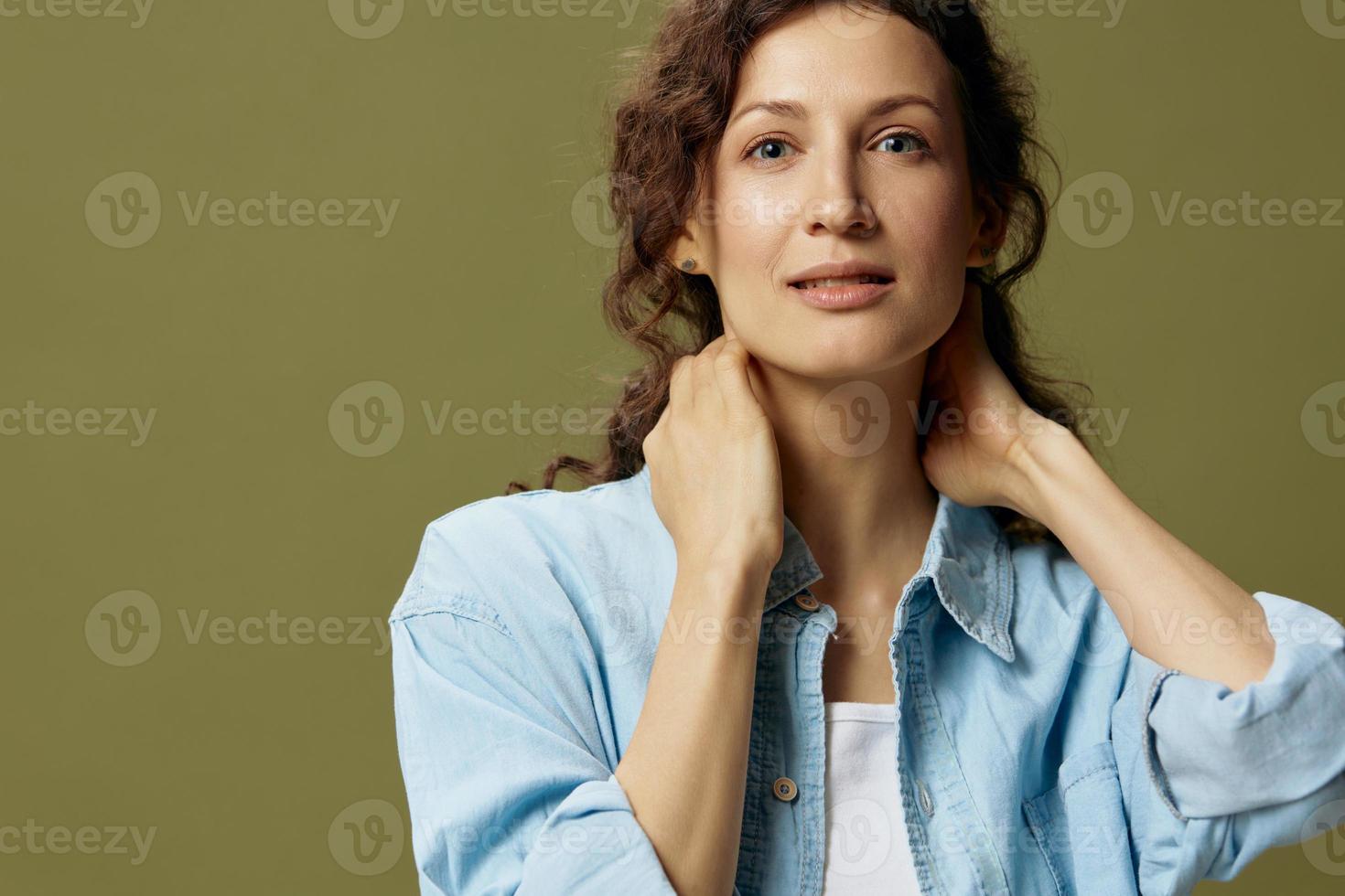 Smiling adorable curly beautiful female in jeans casual shirt touches neck posing isolated on over olive green pastel background. Being Yourself. People Lifestyle emotions concept. Copy space photo