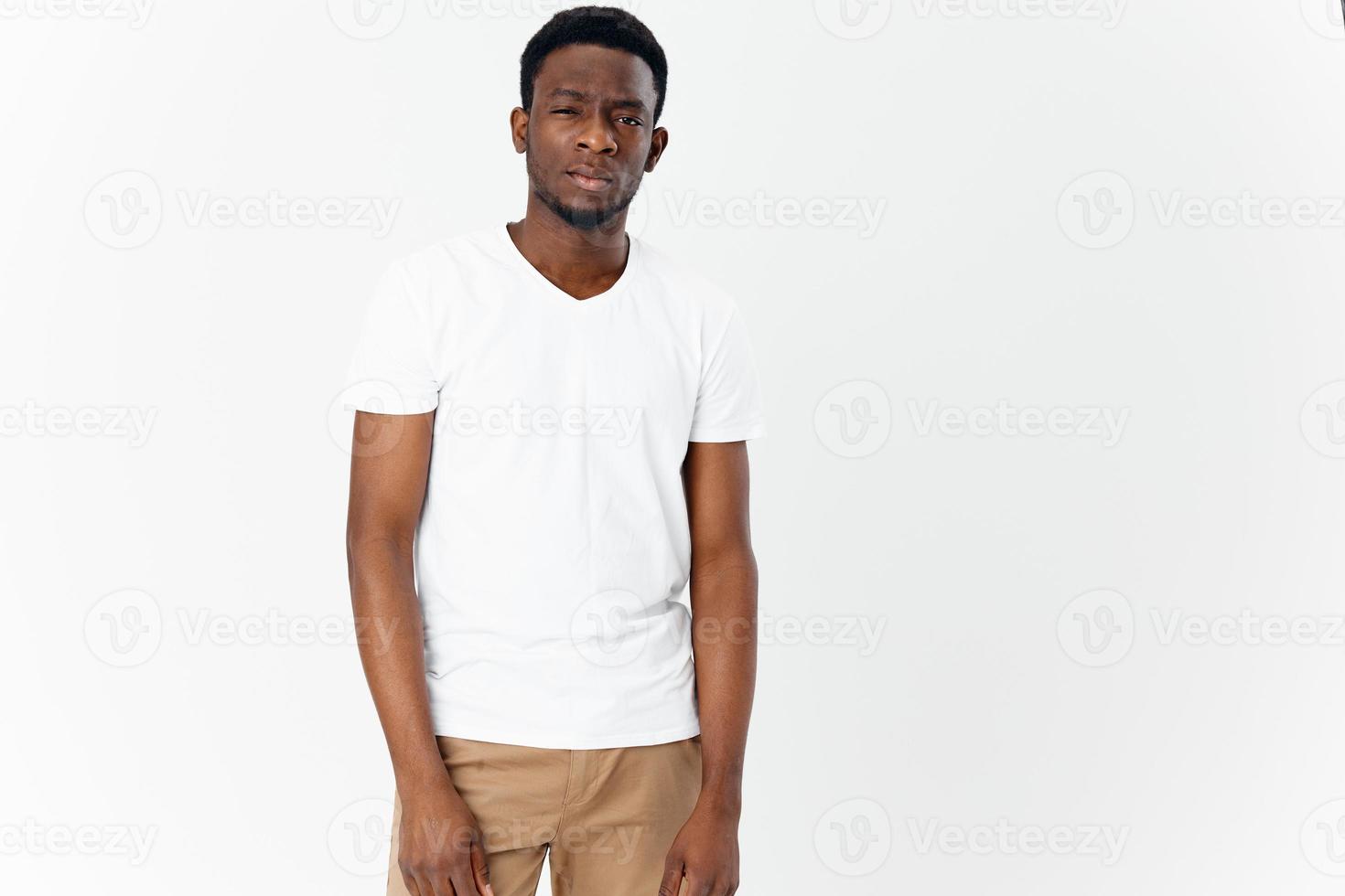 un joven hombre de africano apariencia en beige pantalones y un blanco camiseta mira adelante en un ligero antecedentes foto