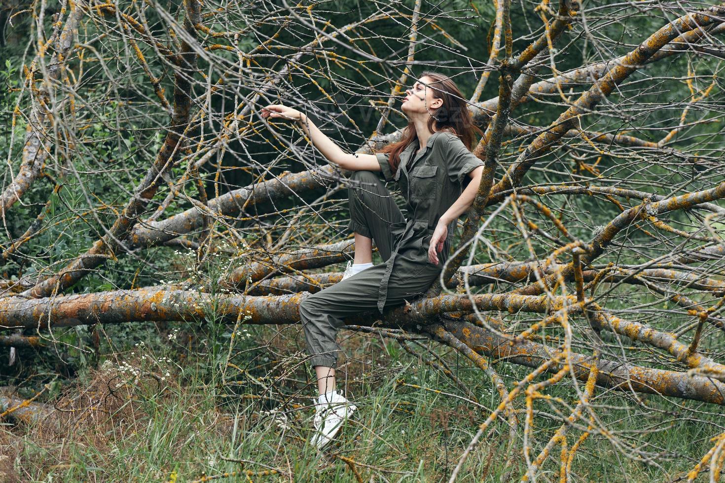 A woman in a green jumpsuit sits on a broken tree in sneakers photo