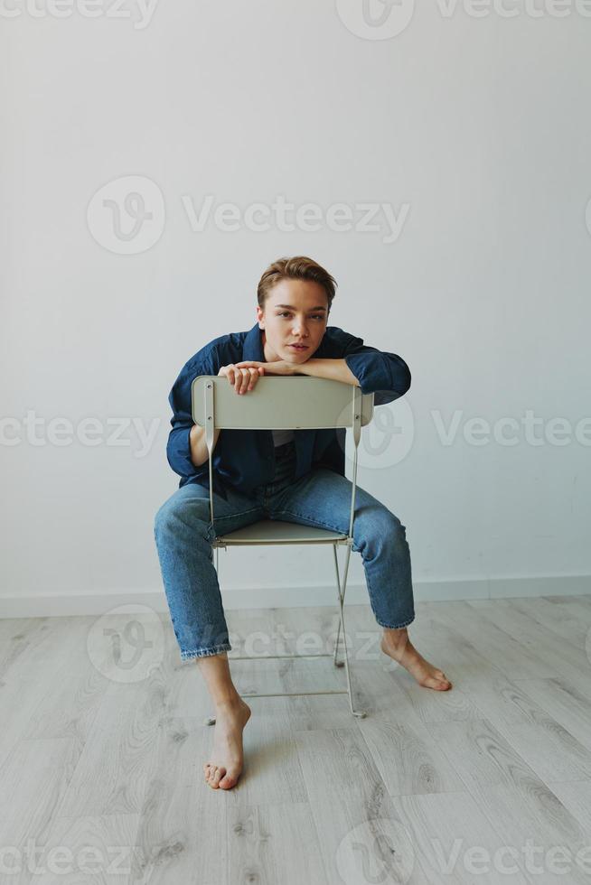 A young woman sitting in a chair at home smiling with teeth with a short haircut in jeans and a denim shirt on a white background. Girl natural poses with no filters photo