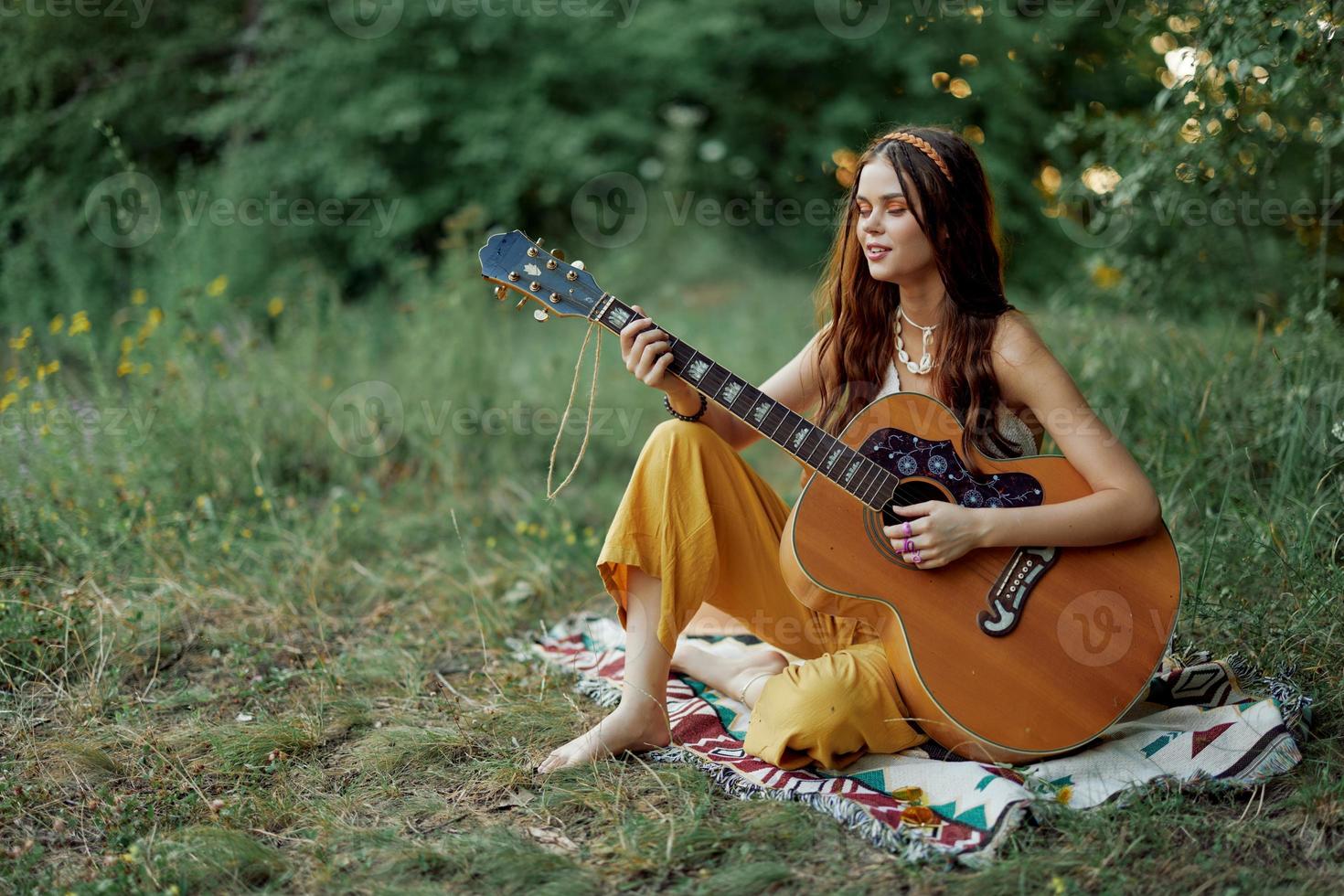 joven hippie mujer sentado al aire libre en un tartán y jugando su guitarra en el orilla del río dentro el puesta de sol foto