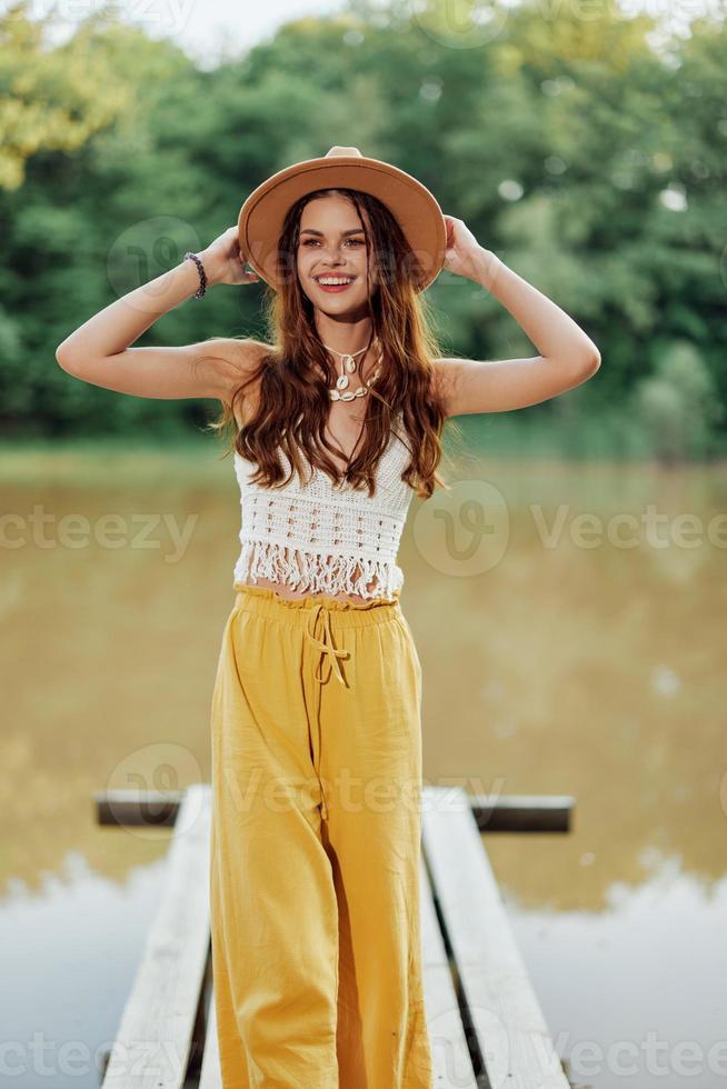 A young woman in a hippie look and eco clothing travels outdoors by the lake wearing a hat and yellow pants in the fall photo