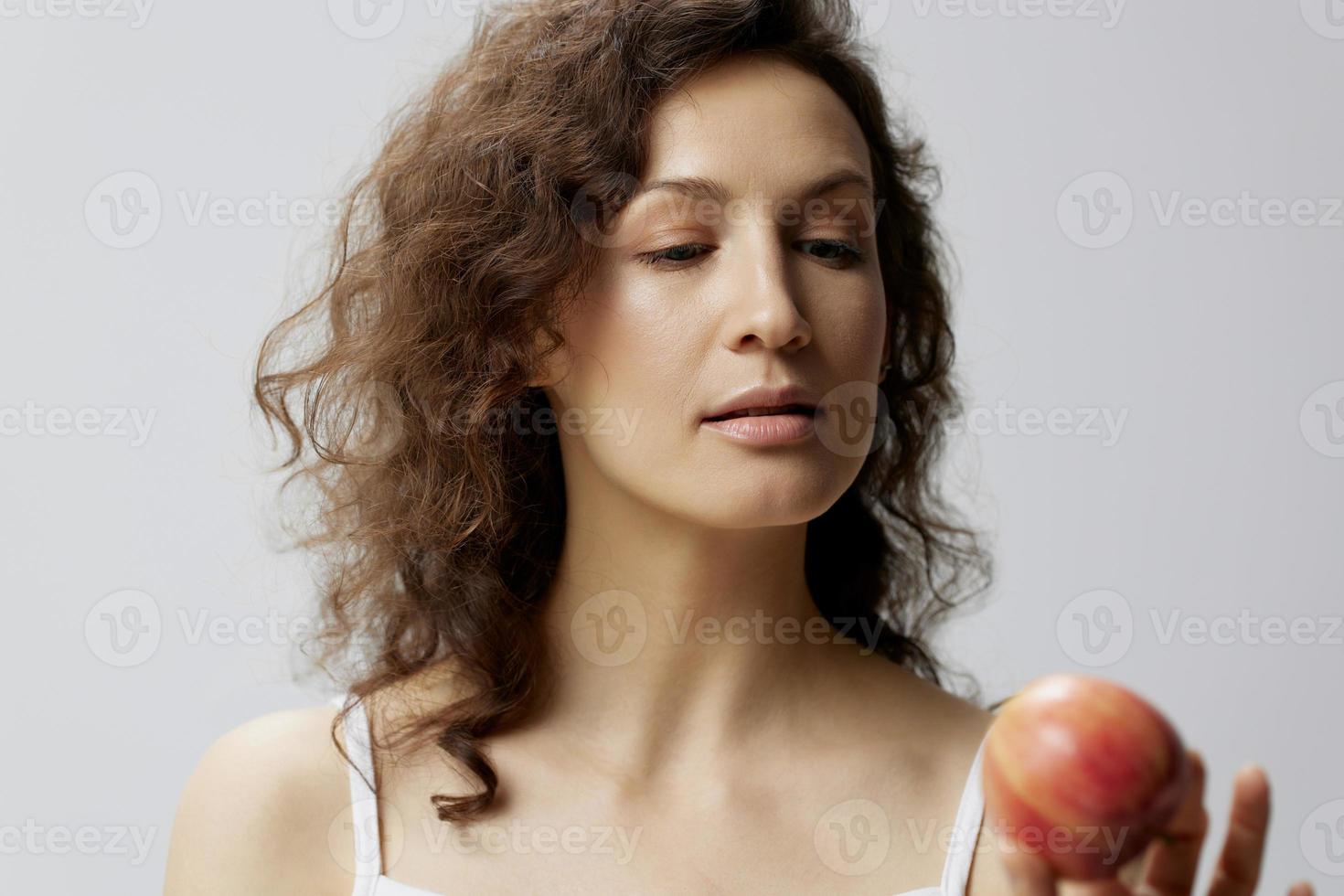 sonriente pensativo linda Rizado hermosa mujer en básico blanco camiseta mira a manzana disfrutar sano comida posando aislado en terminado blanco antecedentes. natural Respetuoso del medio ambiente productos concepto. Copiar espacio foto