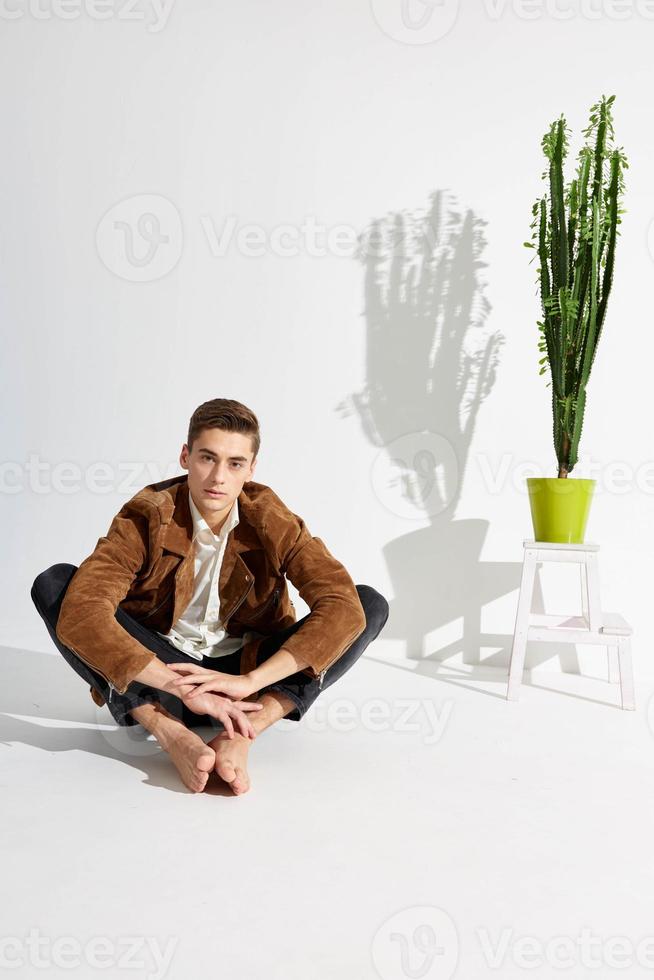 fashionable man in a jacket and trousers sits barefoot on the floor near a flower in a pot photo