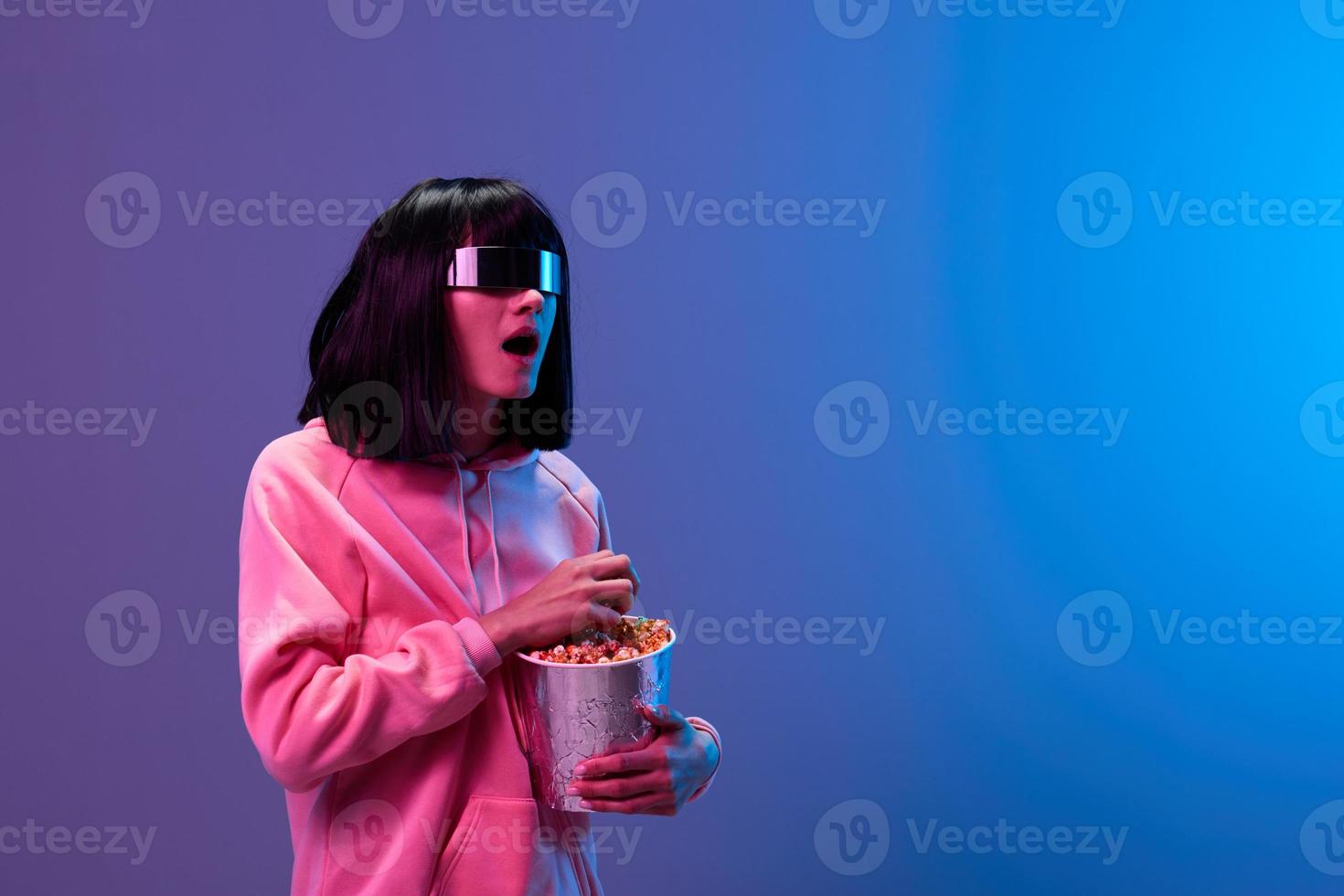 Shocked adorable brunet woman in pink hoodie trendy specular sunglasses with popcorn open mouth look aside posing isolated in blue violet color light background. Neon party Cinema concept. Copy space photo