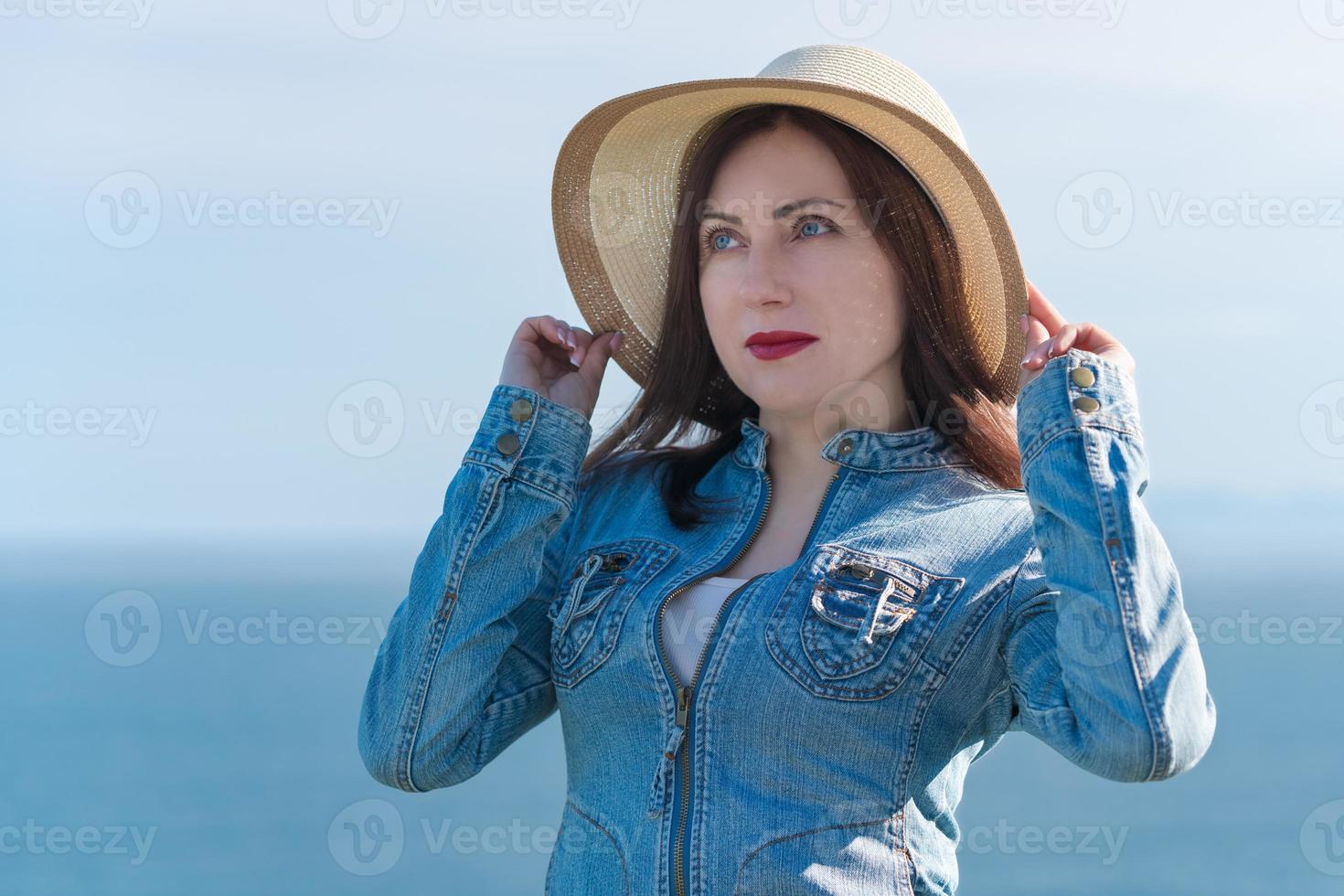 40 year old hipster woman raised hands and holds straw hat on head, posing against sky and ocean photo