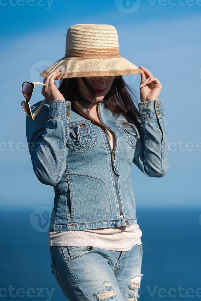 Woman in denim jacket and jeans hiding with straw hat over his face on background of sky and ocean photo