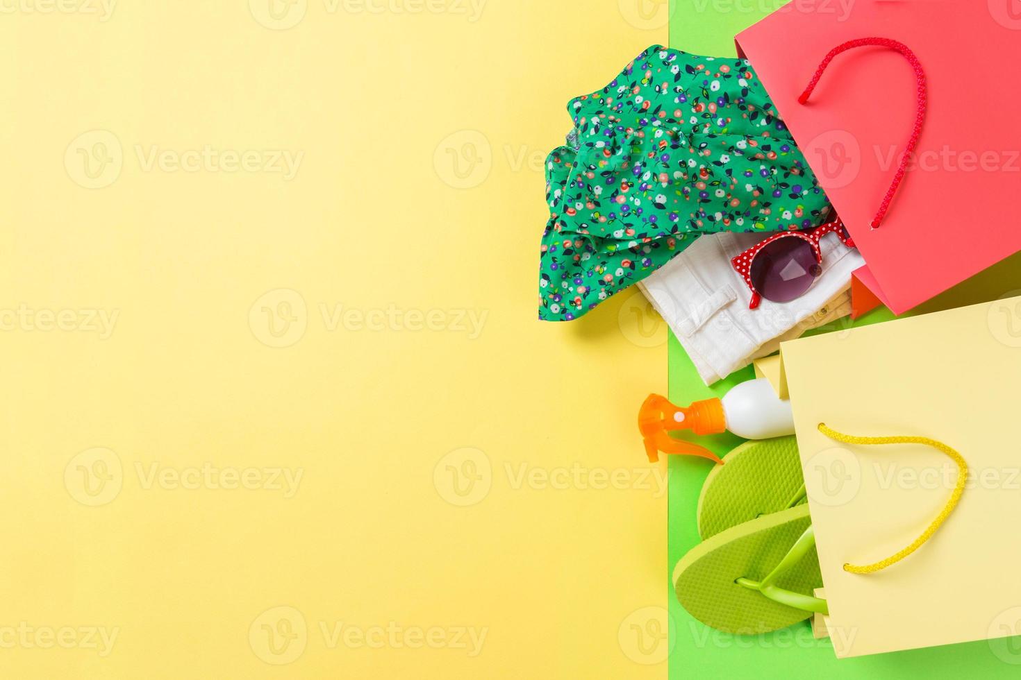 Summer concept of colourful shopping bags full of clothes. Gift bags with T-shirt, denim shorts, flip flops and bottle of sunscreen. Summer wishlist concept. Top view copy space on yellow background photo