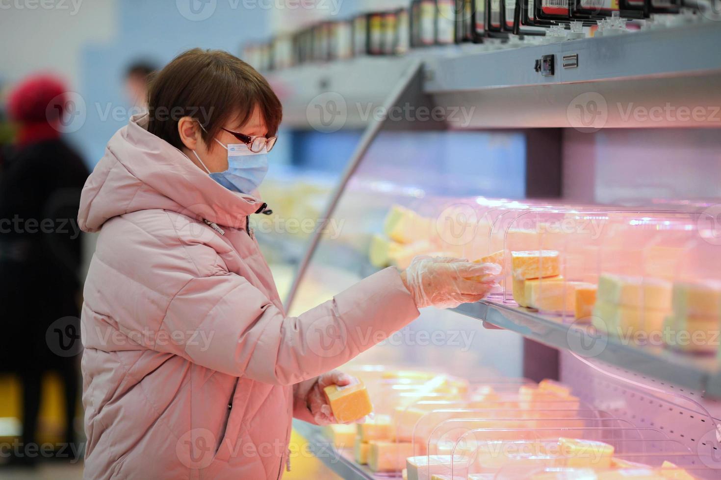 An adult woman in a medical mask and glasses examines a label of cheese in a grocery store. photo