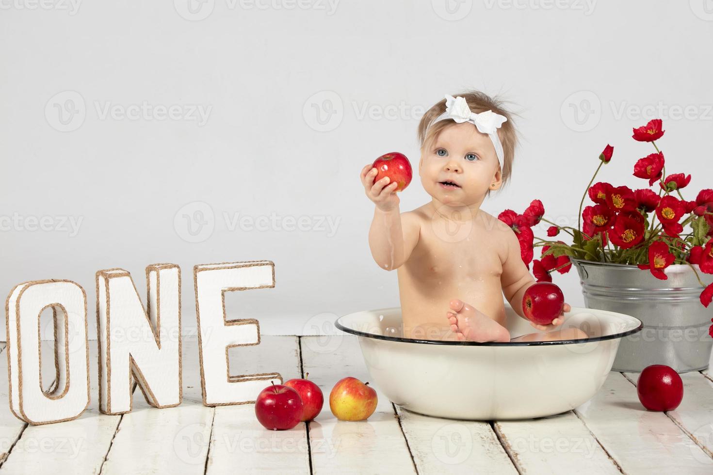 hermosa bebé se baña en un cuenca y come un rojo manzana. pequeño niña en uno año viejo. foto