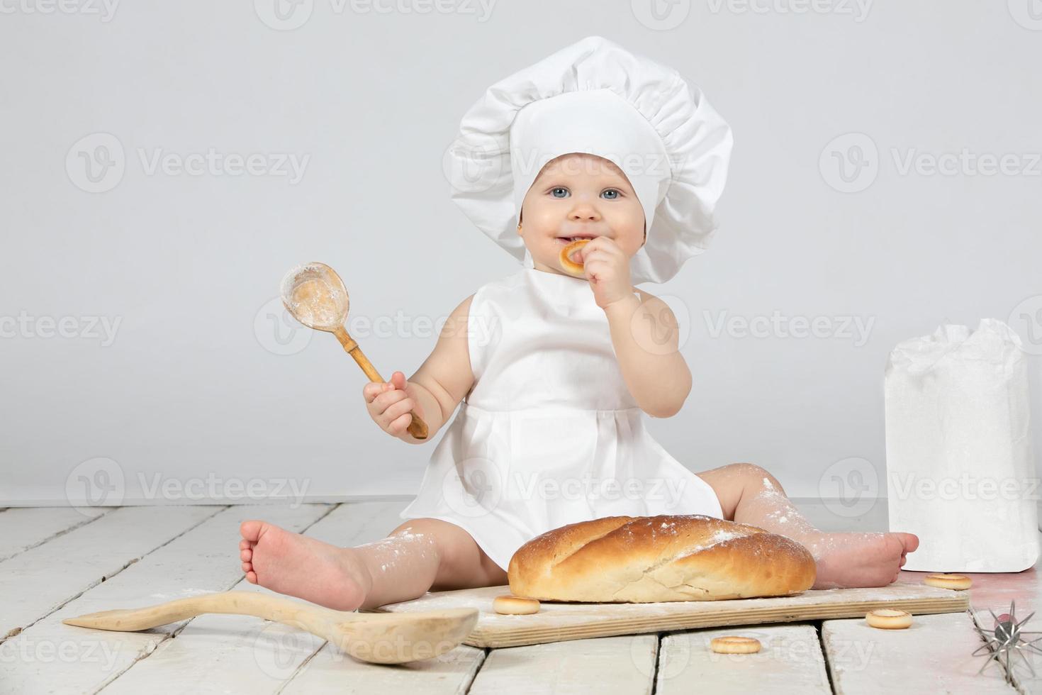 Child cook. Beautiful little girl in cook clothes with a long loaf and a wooden spoon in flour. The child is one year old. Funny baby. Little chef. The concept of baking bakery products. photo