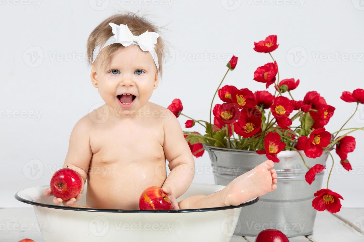 A child bathes in a basin. Beautiful baby with apples on a background of red flowers. Girl in one year. Beautiful child. photo