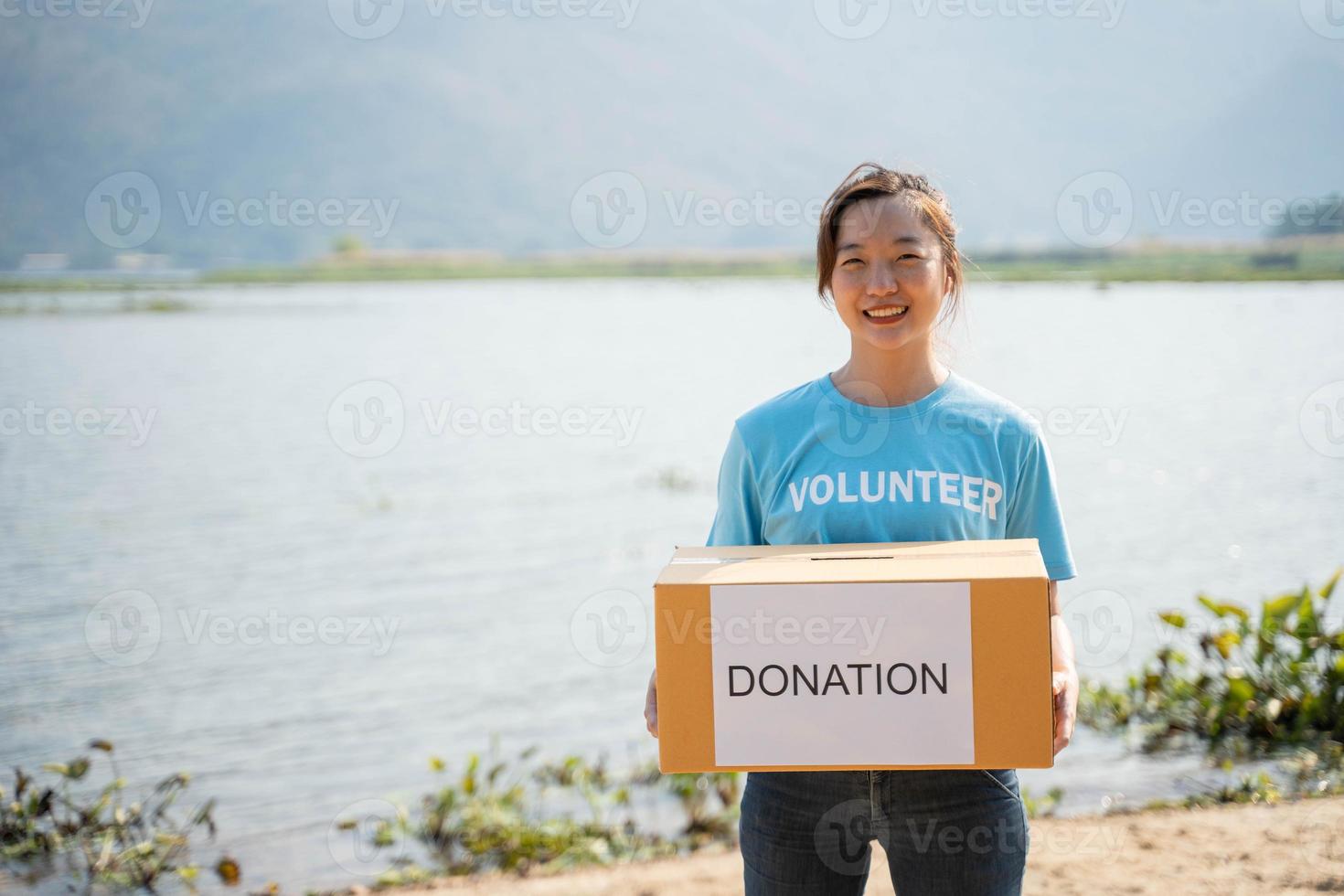 Portrait of young woman wearing volunteer t-shirt,  holding donations box and looking at camera, standing on river background photo