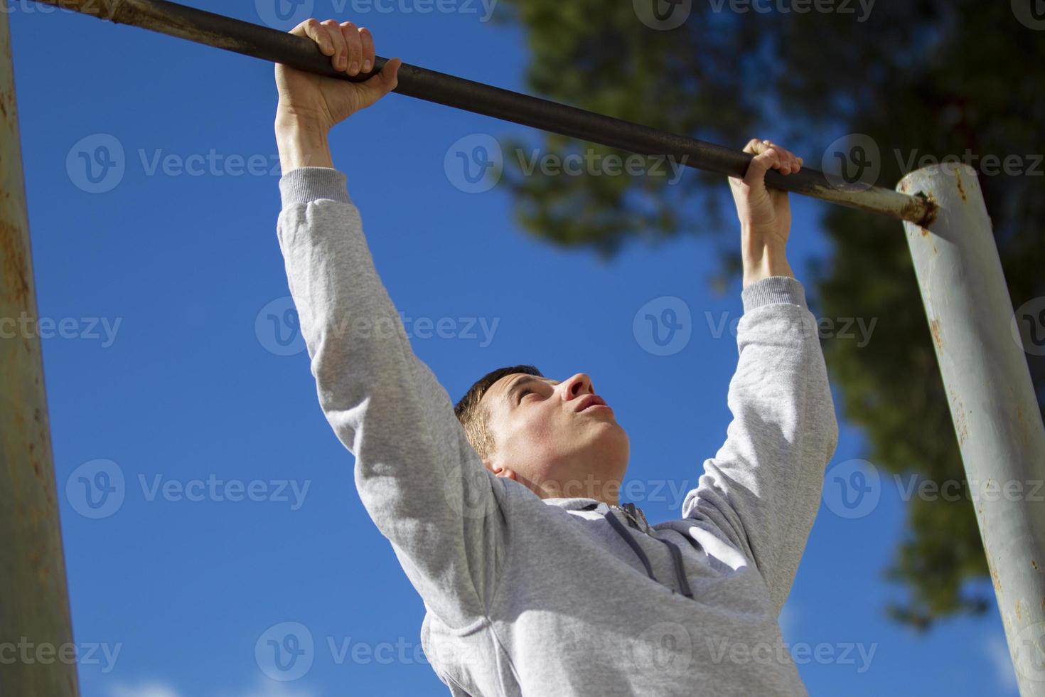 The boy pulls himself up on the horizontal bar on the street. A young man does exercises. photo