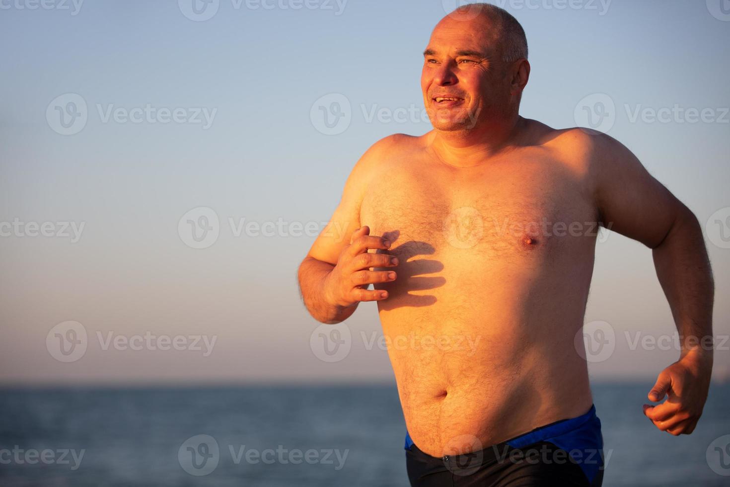 A tanned man runs against the background of the sea. An elderly man of athletic build runs along the coast. photo