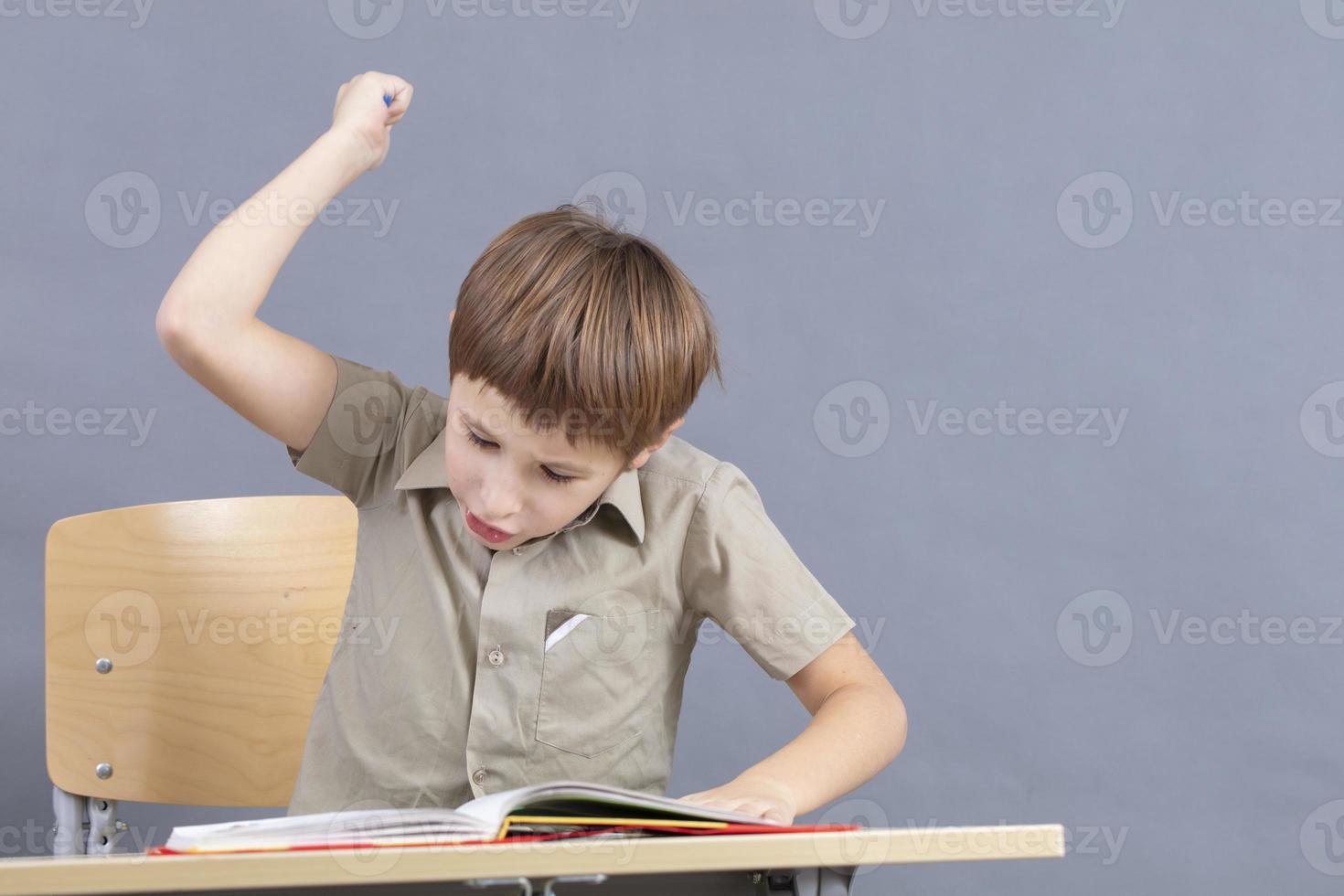 A primary school student is doing homework. The child at the desk with the textbook is angry. The boy does not want to study. A seven-year-old child goes crazy for lessons. photo