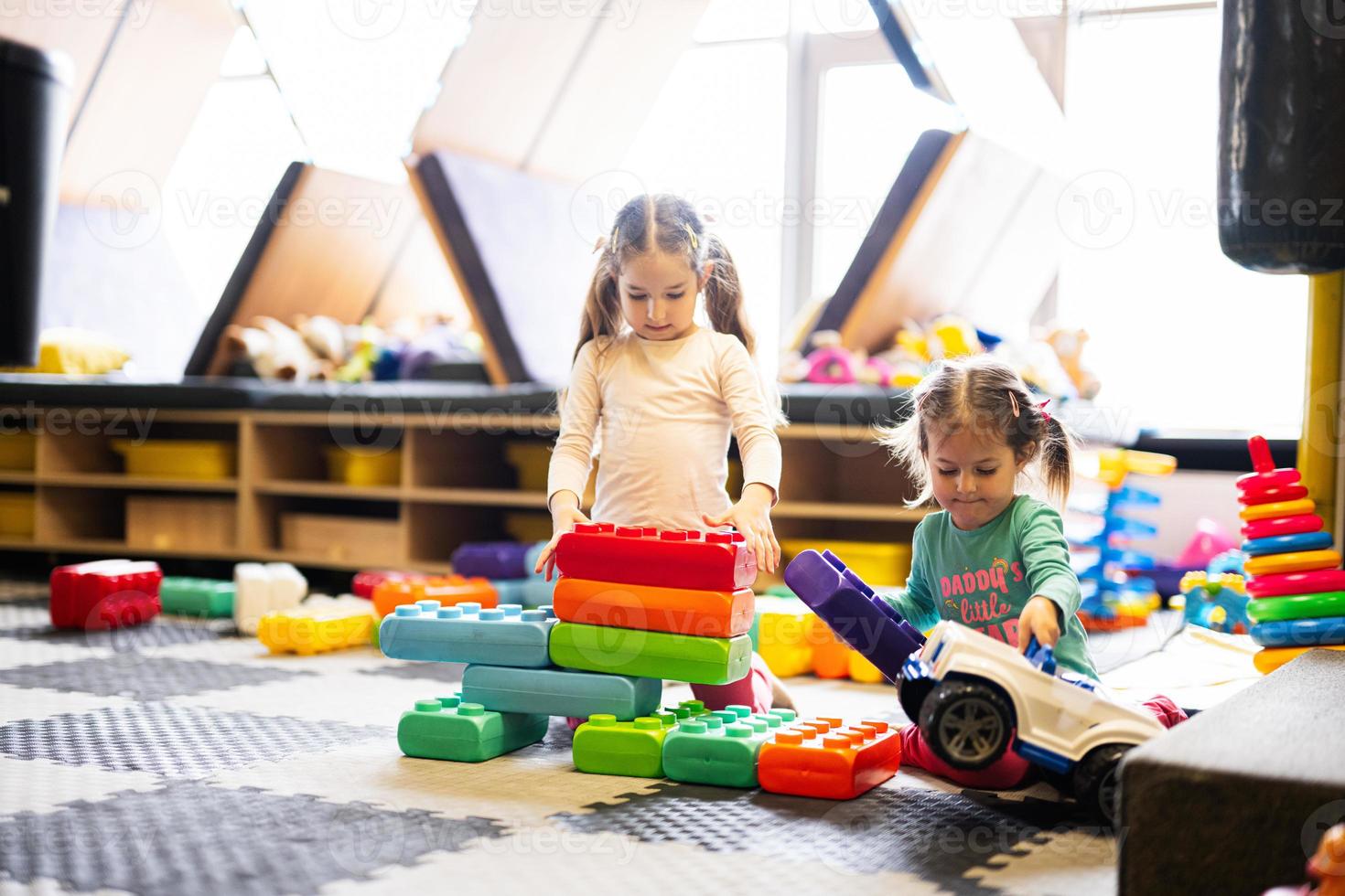 Two sisters playing at kids play center while build with colored plastic blocks. photo