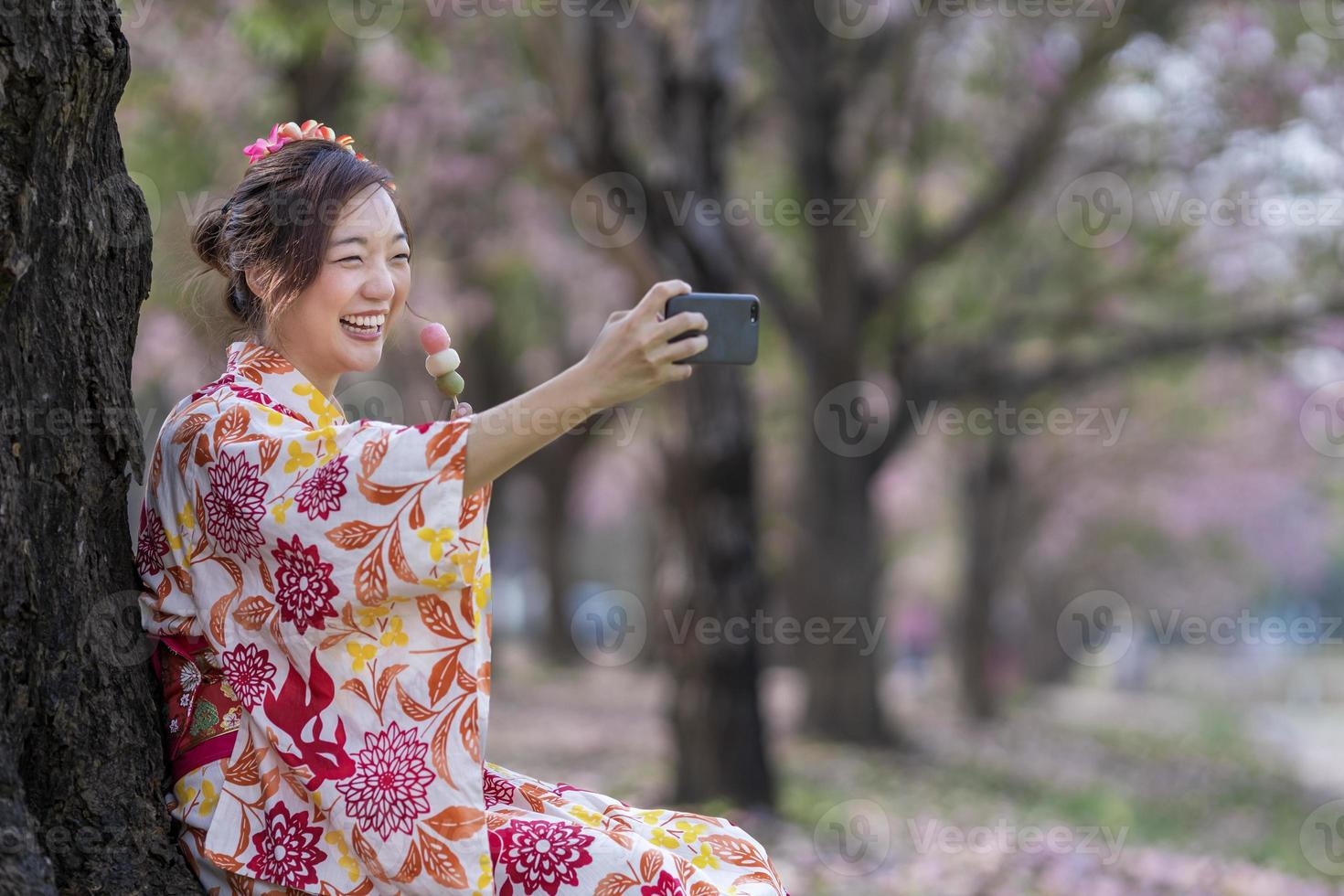 japonés mujer en tradicional kimono tomando selfie hanami dango gesto mientras caminando en el parque a Cereza florecer árbol durante primavera sakura festival concepto foto