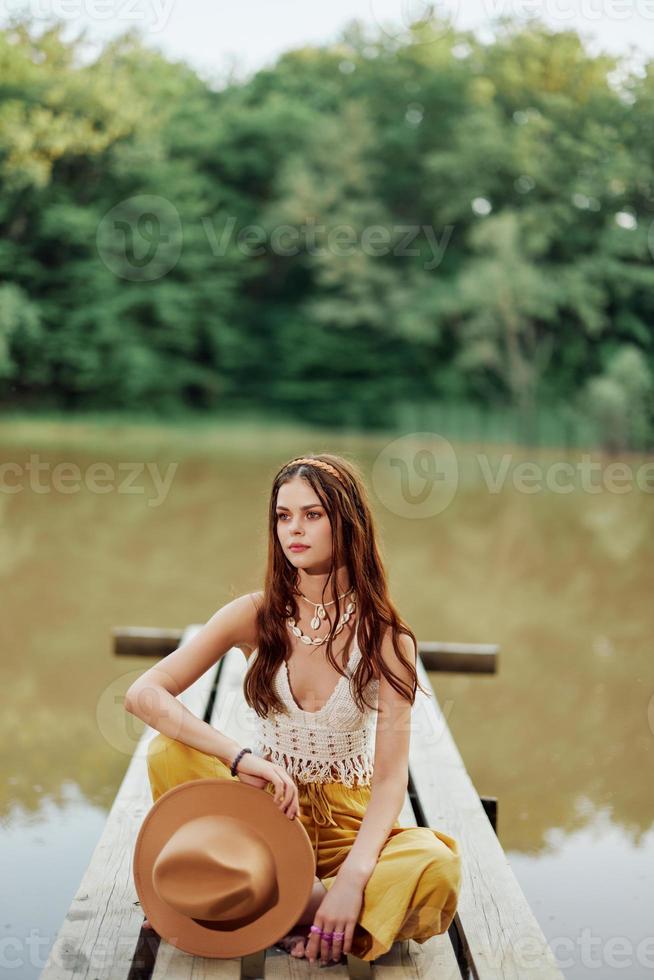 un joven mujer sonriente en un imagen de un hippie y eco-ropa sentado al aire libre en un puente por un lago vistiendo un sombrero y amarillo pantalones en el verano puesta de sol foto