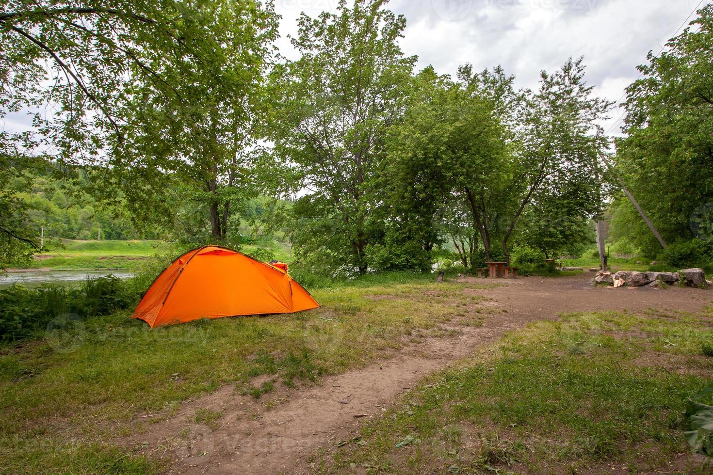 An orange double tent stands in a clearing in a forest by the river. The sky is gray with clouds. photo