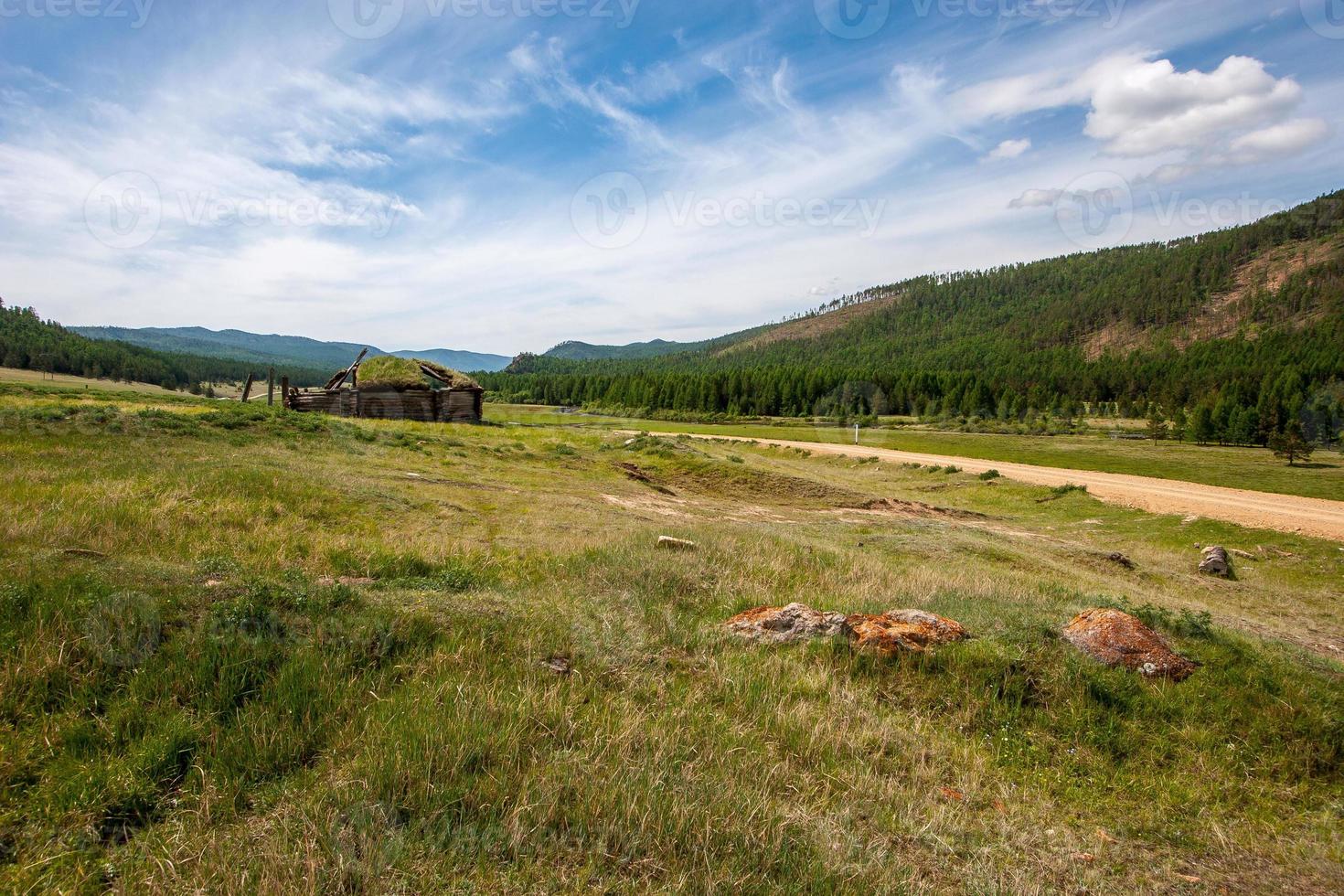 A dirt road between the hills and a ruined old yurt on the grass. Overgrown with grass hut. Clouds on the blue sky. Sunny. Horizontal. photo