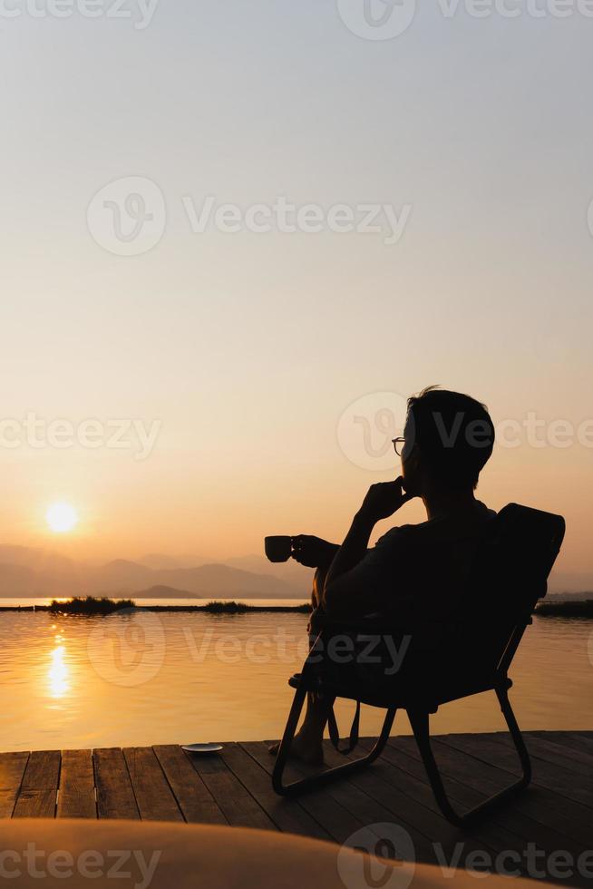 silueta turista hombre sentar en un silla participación taza de café viendo amanecer. foto