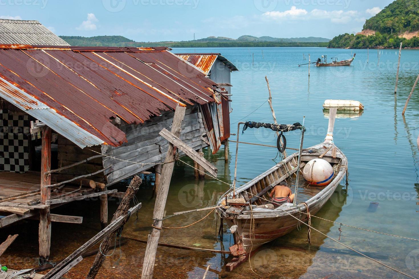Thai fishing long boat next to the fisherman's hut standing on stilts in the water. Rusty Shack Roof. A fisherman pours water from a boat. Big buoy in the boat. photo