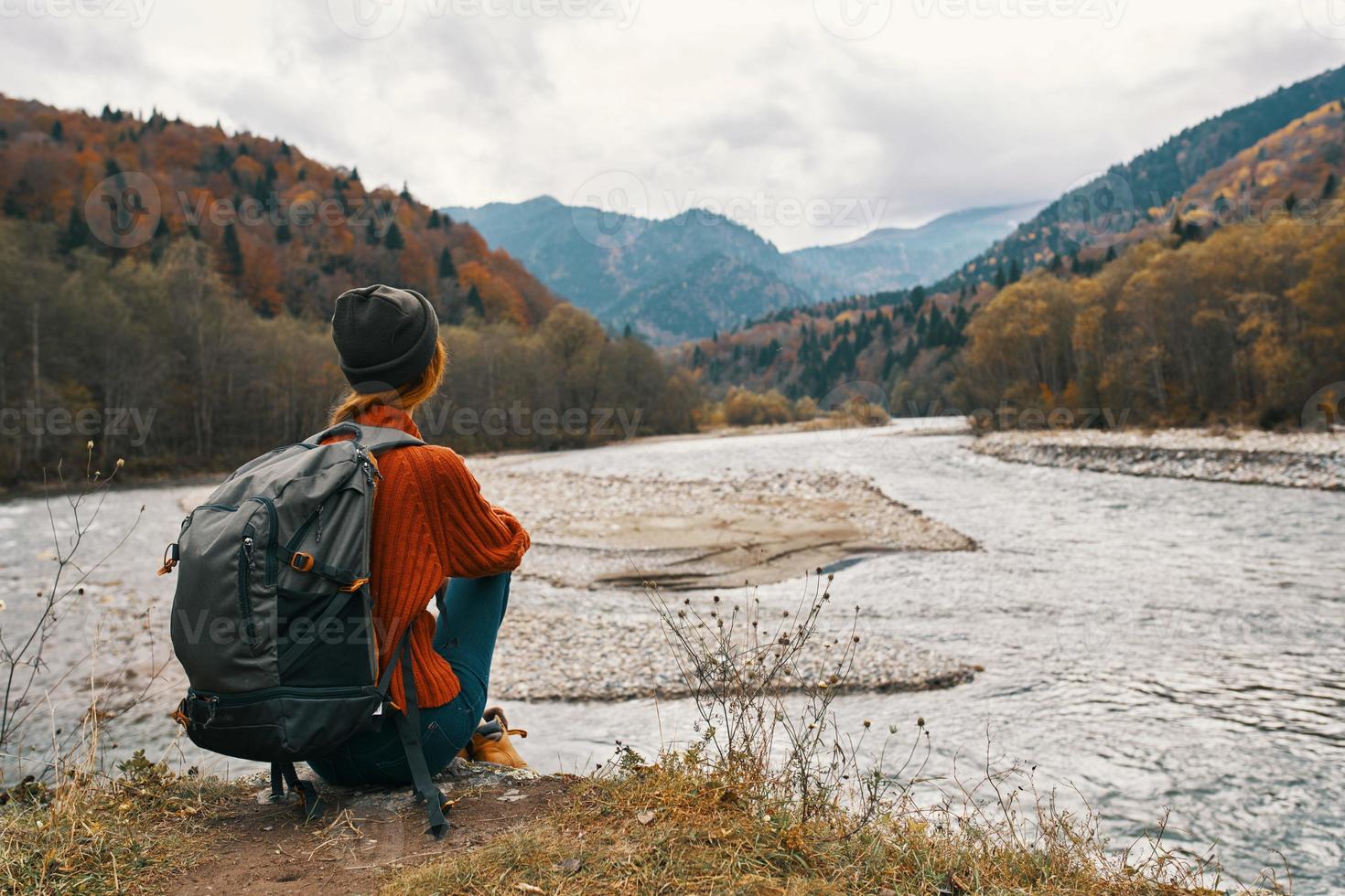 woman in the mountains in autumn with a backpack Sits on the river bank and looks at the high mountains photo