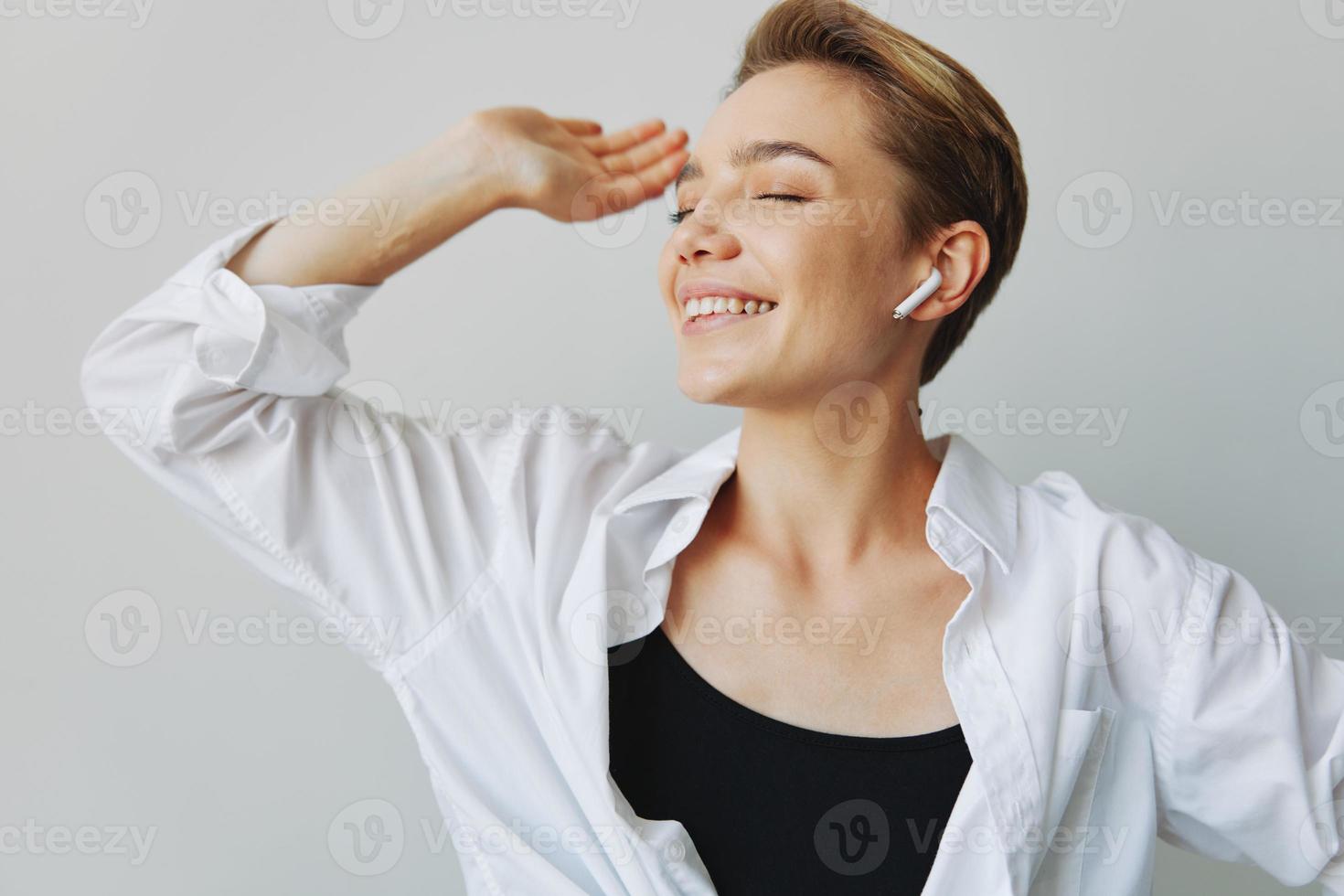 Young woman teenager listening to music with infertile headphones and dancing home, grinning with teeth with a short haircut in a white shirt on a white background. Girl natural poses with no filters photo