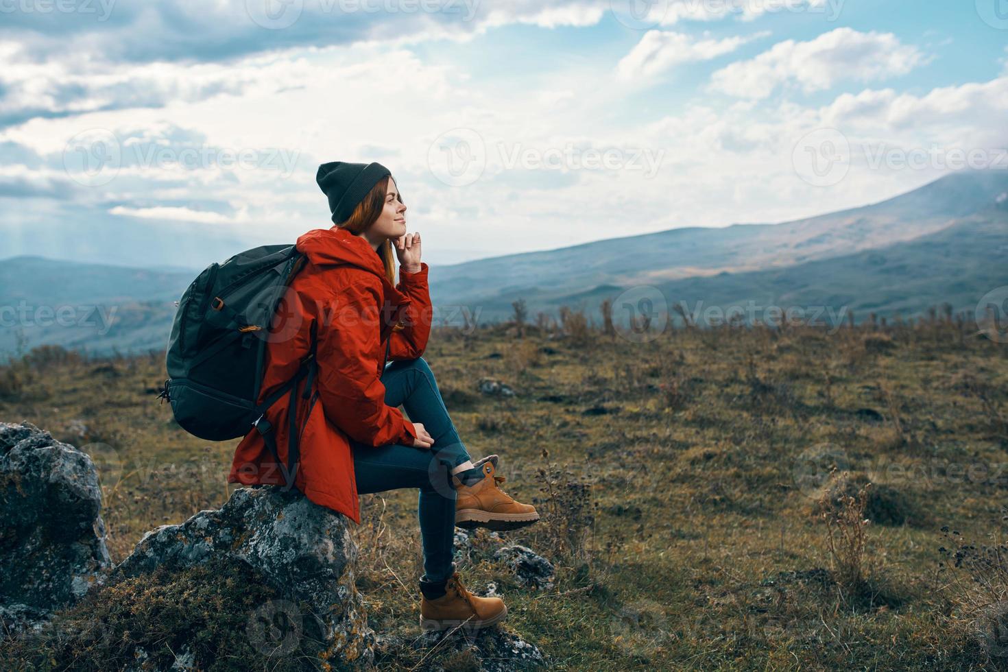 Traveler In a red jacket, hats with a backpack sits on a stone in the mountains in nature photo