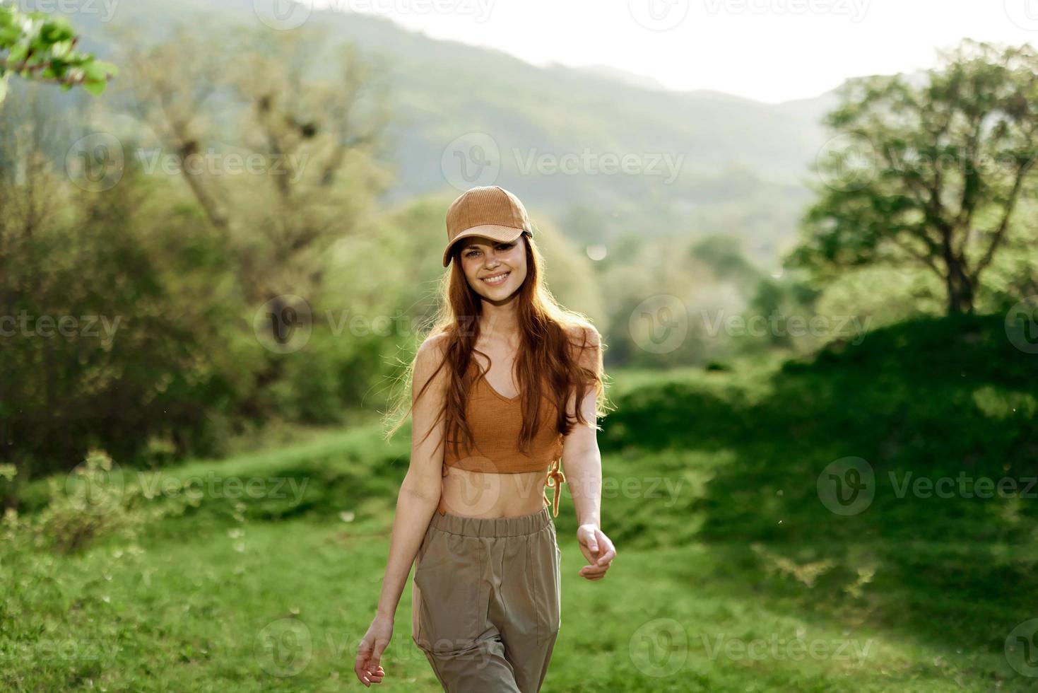 A beautiful woman in sportswear and a cap walks against a green natural landscape and smiles in the sunlight photo