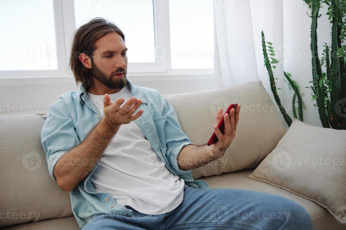 A young man looks angrily and sadly at the phone screen at home sitting on the couch. Bad news and bad luck at work photo