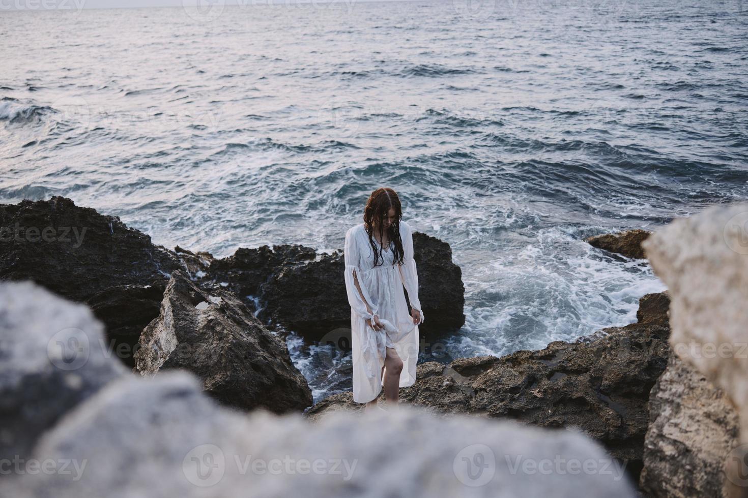 A woman in a white dress stands on rocky stones by the ocean unaltered photo