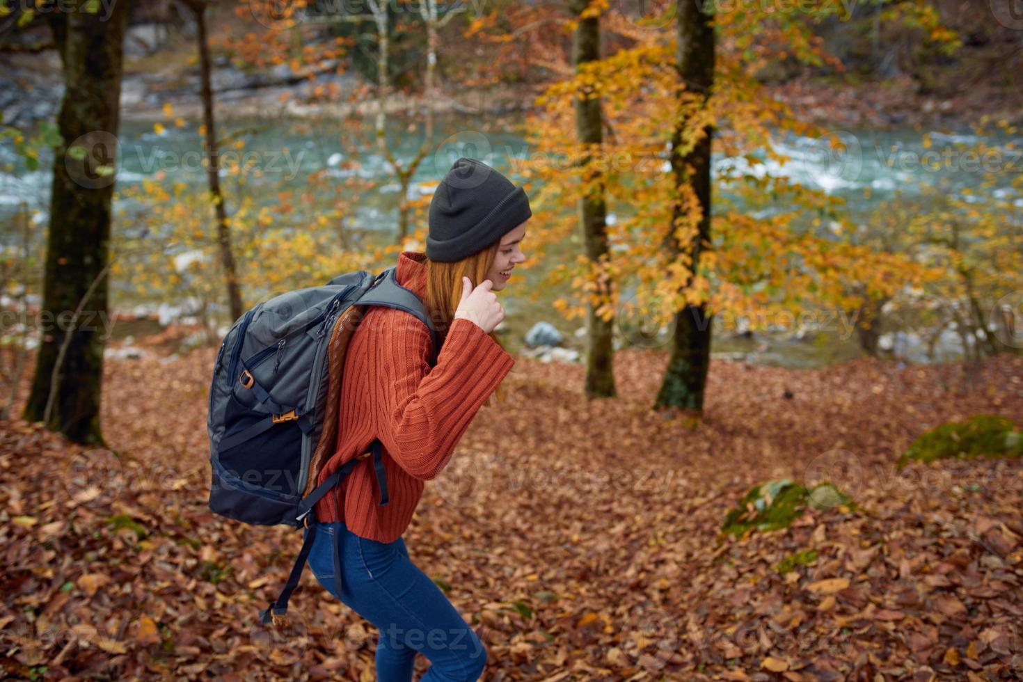 woman in a sweater with a backpack on her back near the river in the mountains and park trees autumn landscape photo