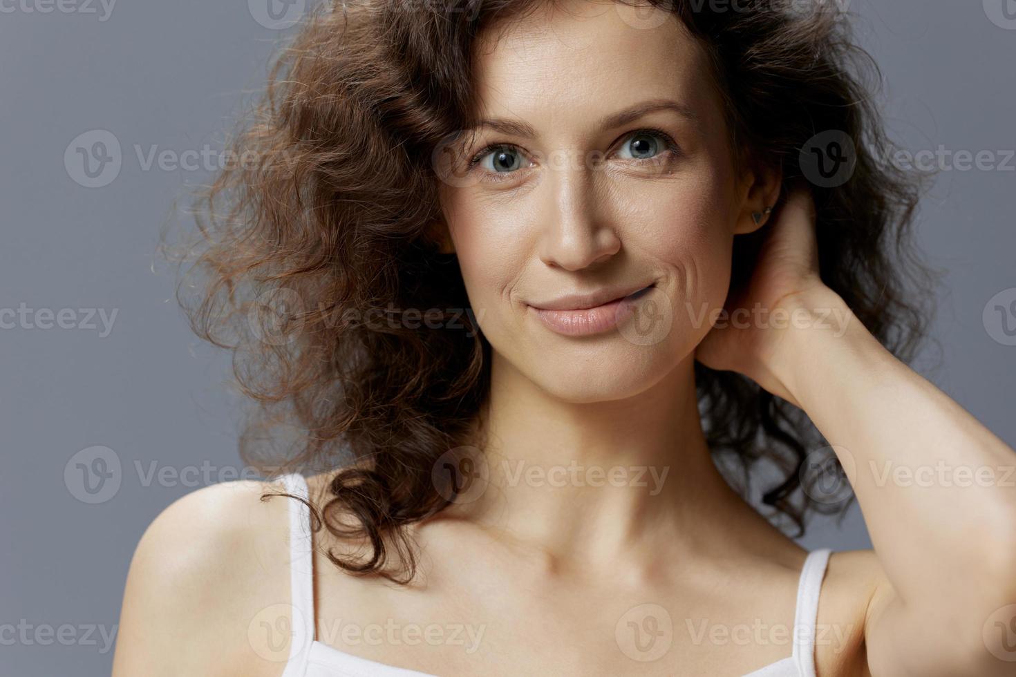 Cheerful shy curly beautiful woman in basic white t-shirt touches neck looks at camera flirting posing isolated on over gray blue background. People Lifestyle Emotions concept. Copy space photo