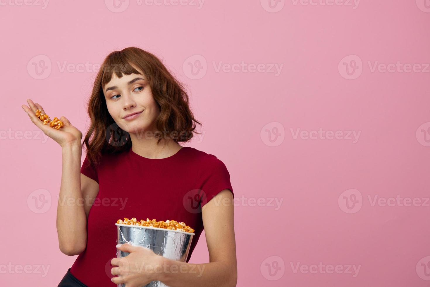 emocionado confuso linda pelirrojo dama en rojo camiseta con palomitas de maiz Listo para película noche posando aislado en terminado rosado estudio antecedentes. Copiar espacio bandera. Moda cine concepto. entretenimiento oferta foto