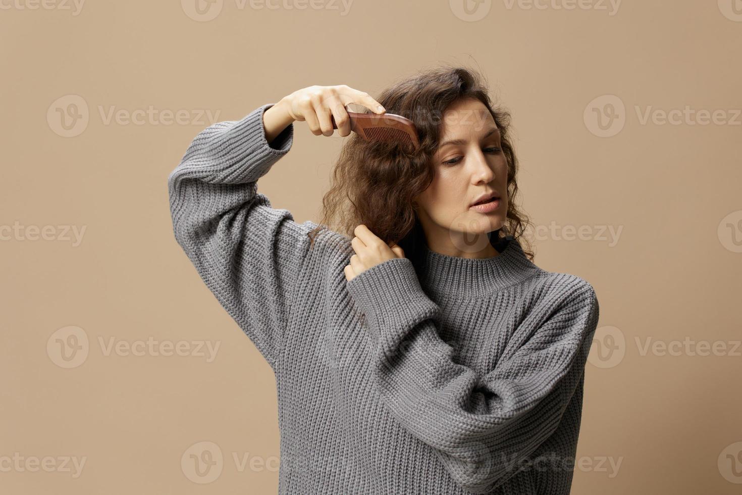 Calm pretty curly beautiful female in gray casual sweater with hairbrush combs hair looks aside posing isolated on over beige pastel background. Problematic unruly damaged hair concept. Copy space photo