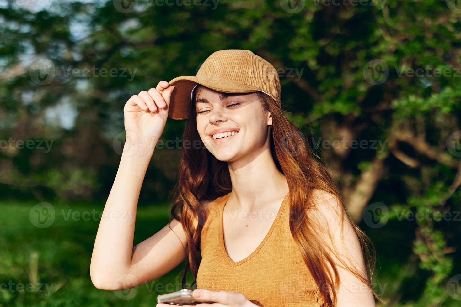 mujer blogger con teléfono en mano en naturaleza en contra un fondo de verdor sonriente en el Brillo Solar vistiendo un gorra después hacer ejercicio foto