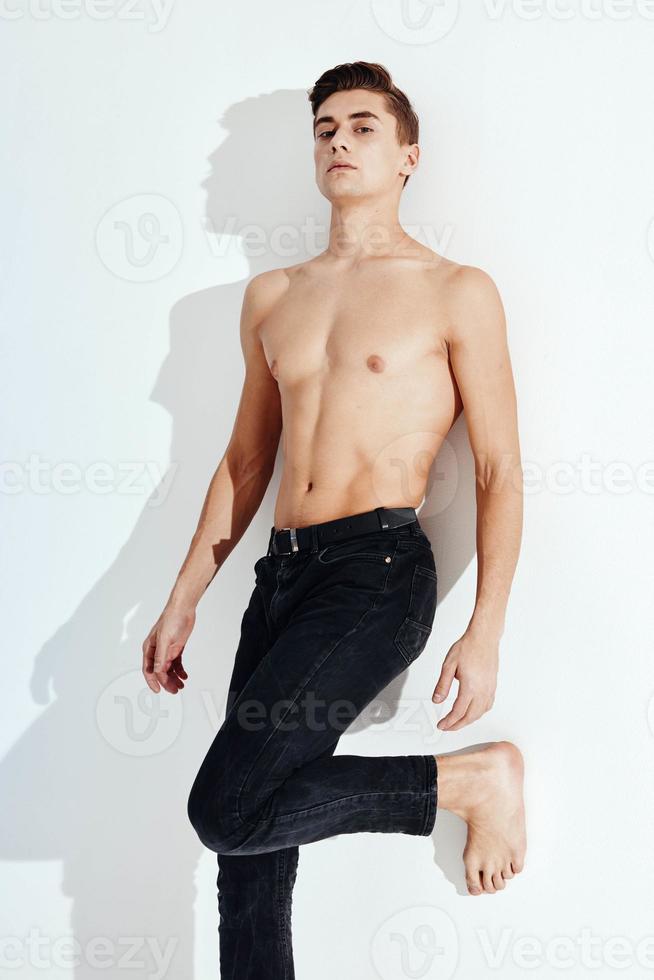 A handsome man in black trousers touches his legs with his hand on a light background near the wall photo