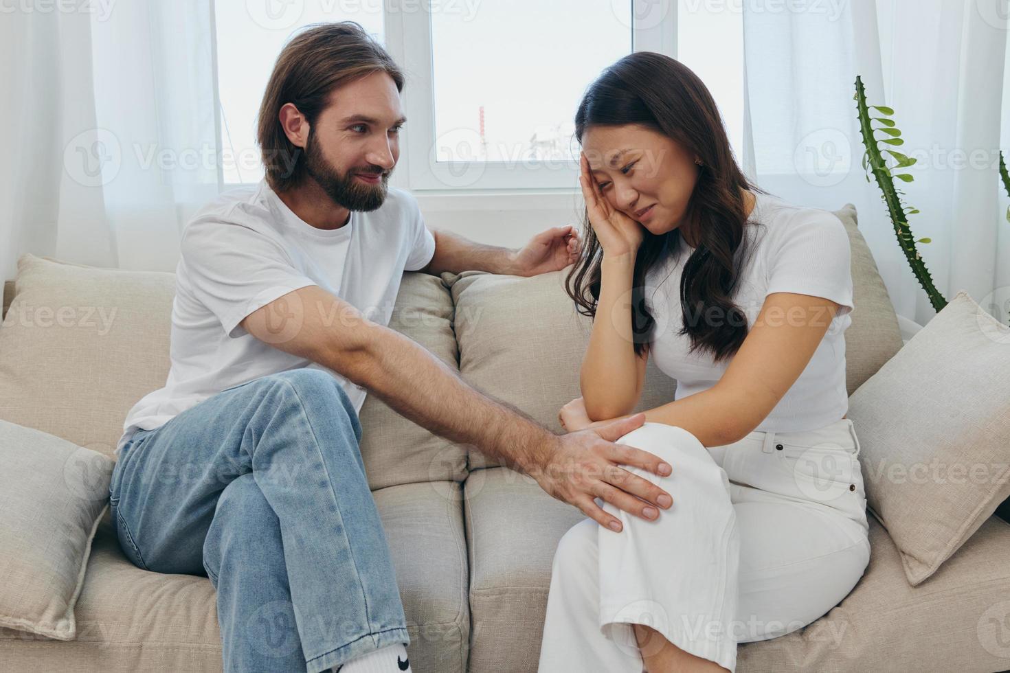 A sad Asian woman talks to a man in tears at home sitting on the couch. Young couple of different nationalities and conflict of interest in a couple photo