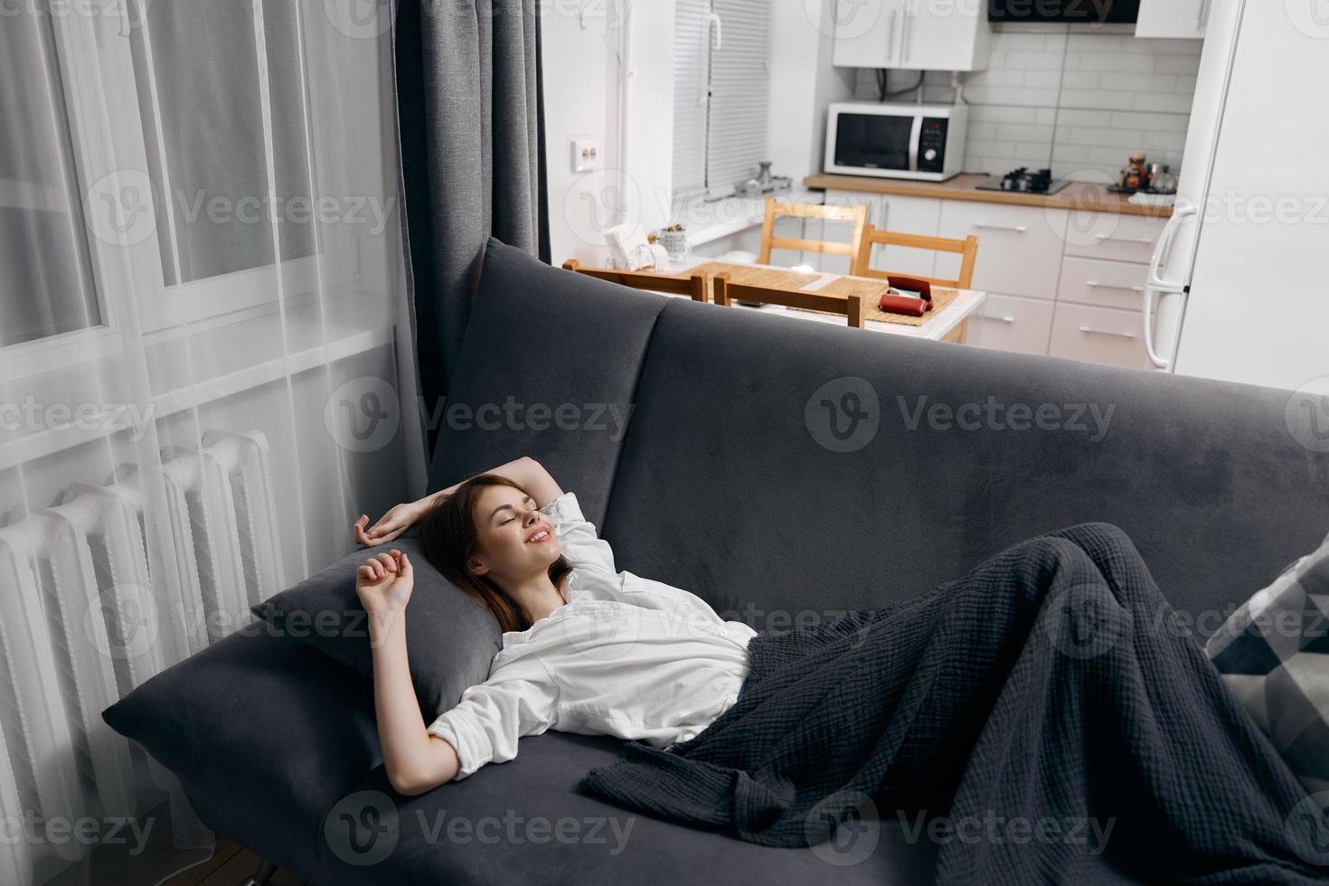 woman covered with a blanket is lying on the sofa and looking up the window in the background photo