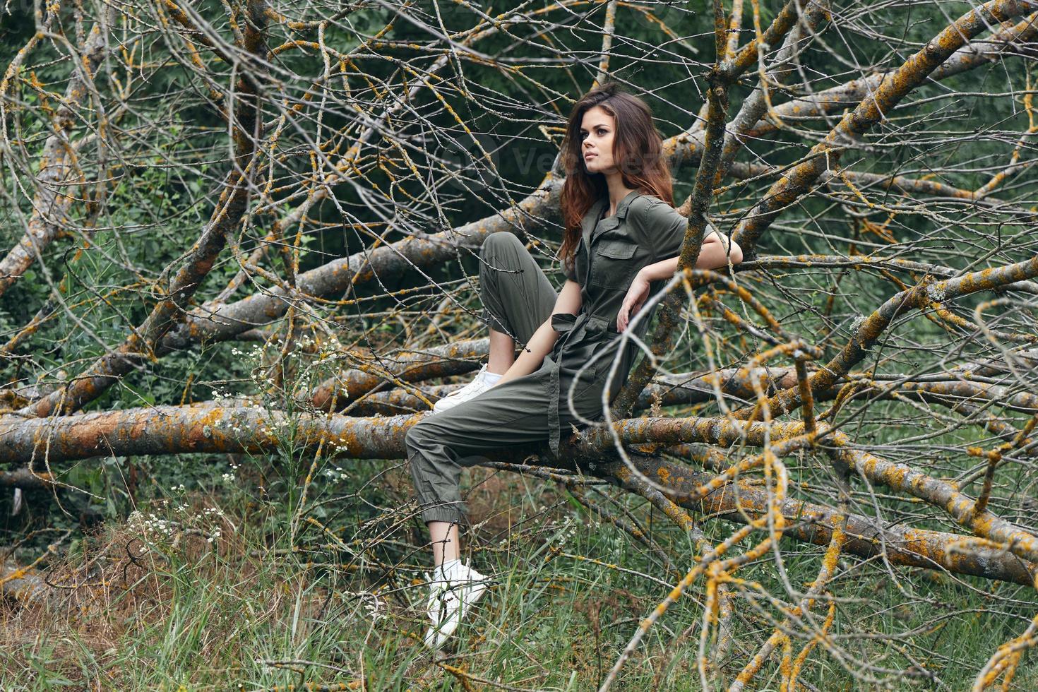 A brunette woman in sneakers and overalls sits on a branch of a broken tree photo